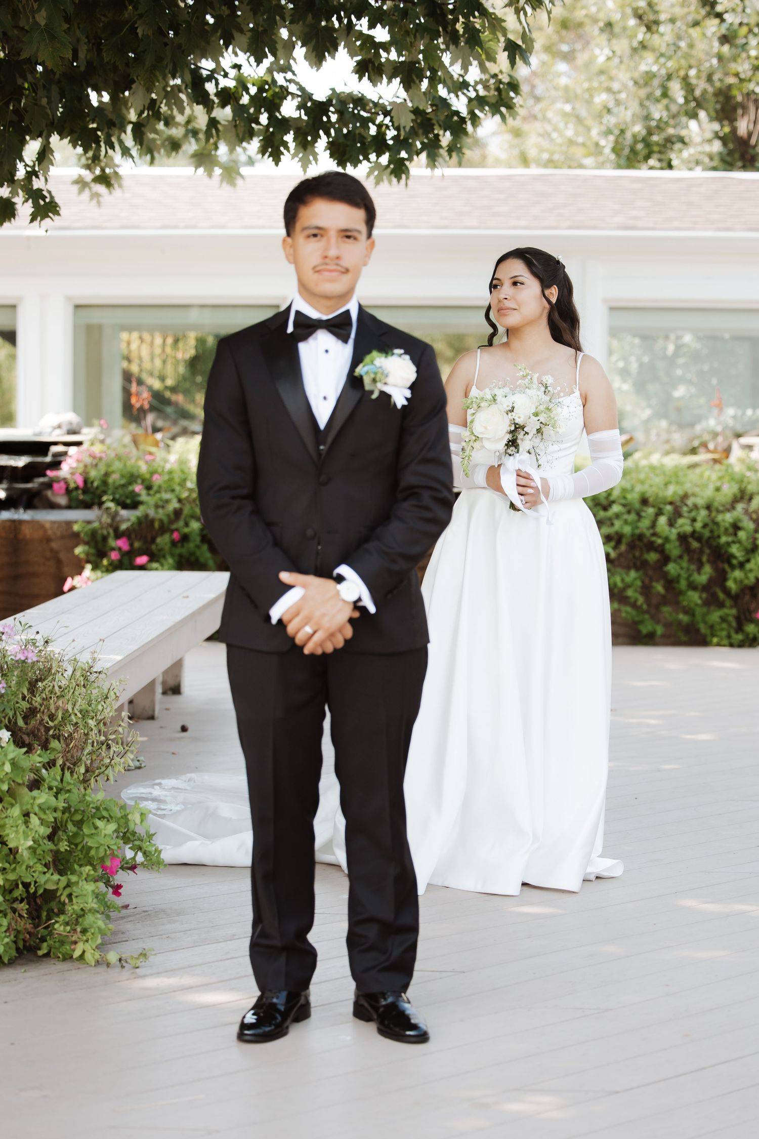 Groom in black tuxedo stands before bride in white wedding dress holding white bouquet at outdoor venue.