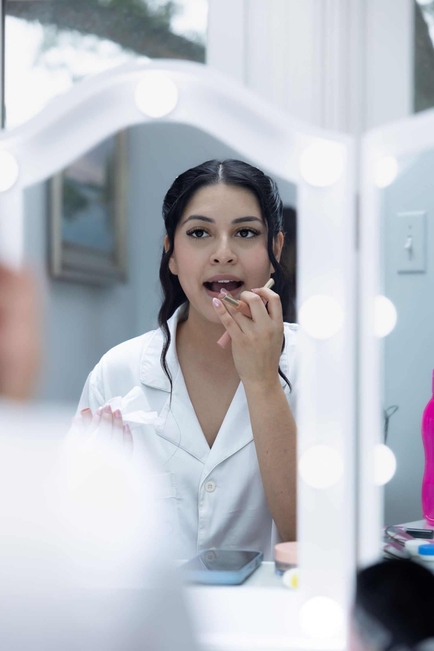 Bride applying lipstick at illuminated vanity mirror during wedding day preparations.