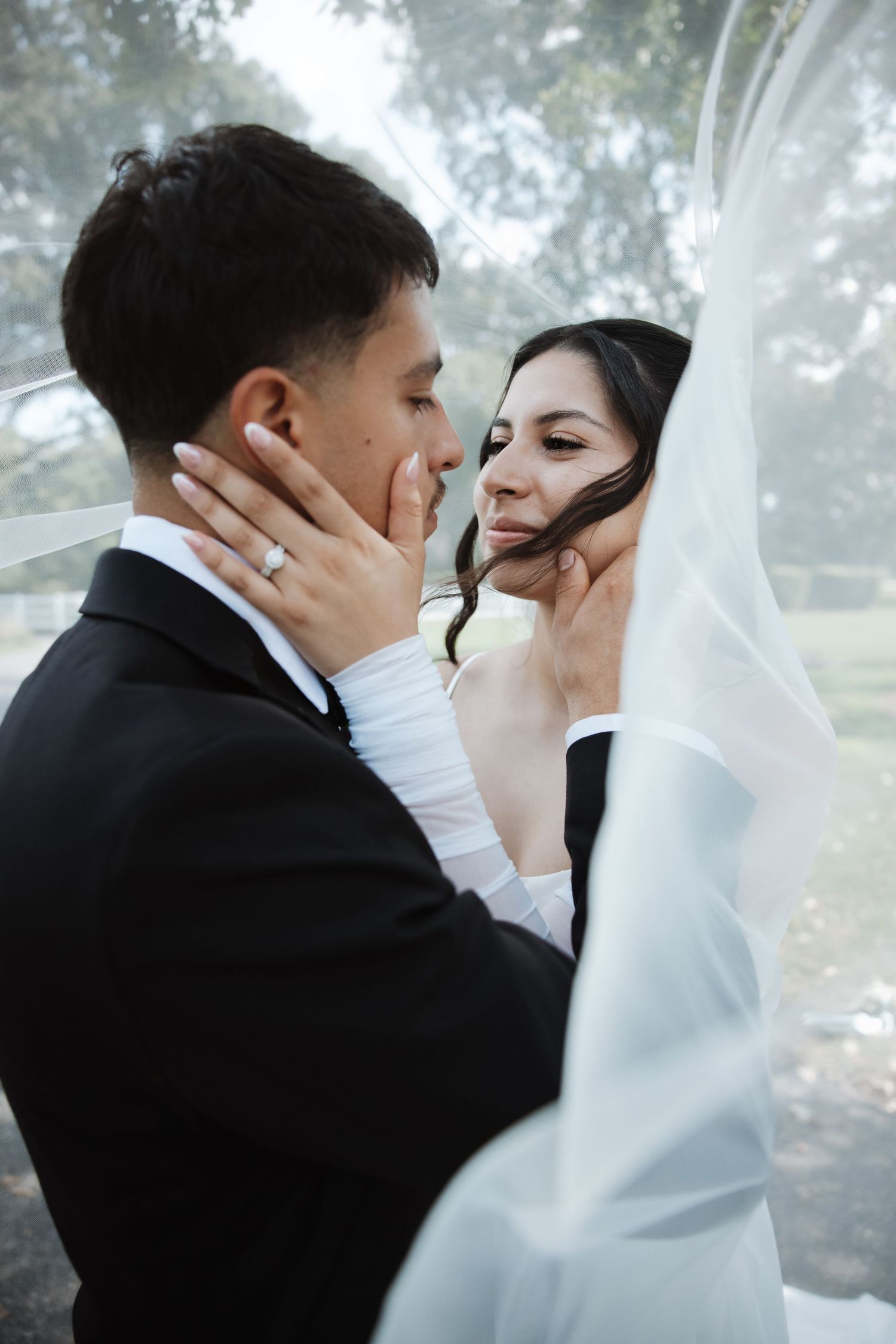 Bride and groom embrace under a billowing white wedding veil outdoors, sharing a tender romantic moment.