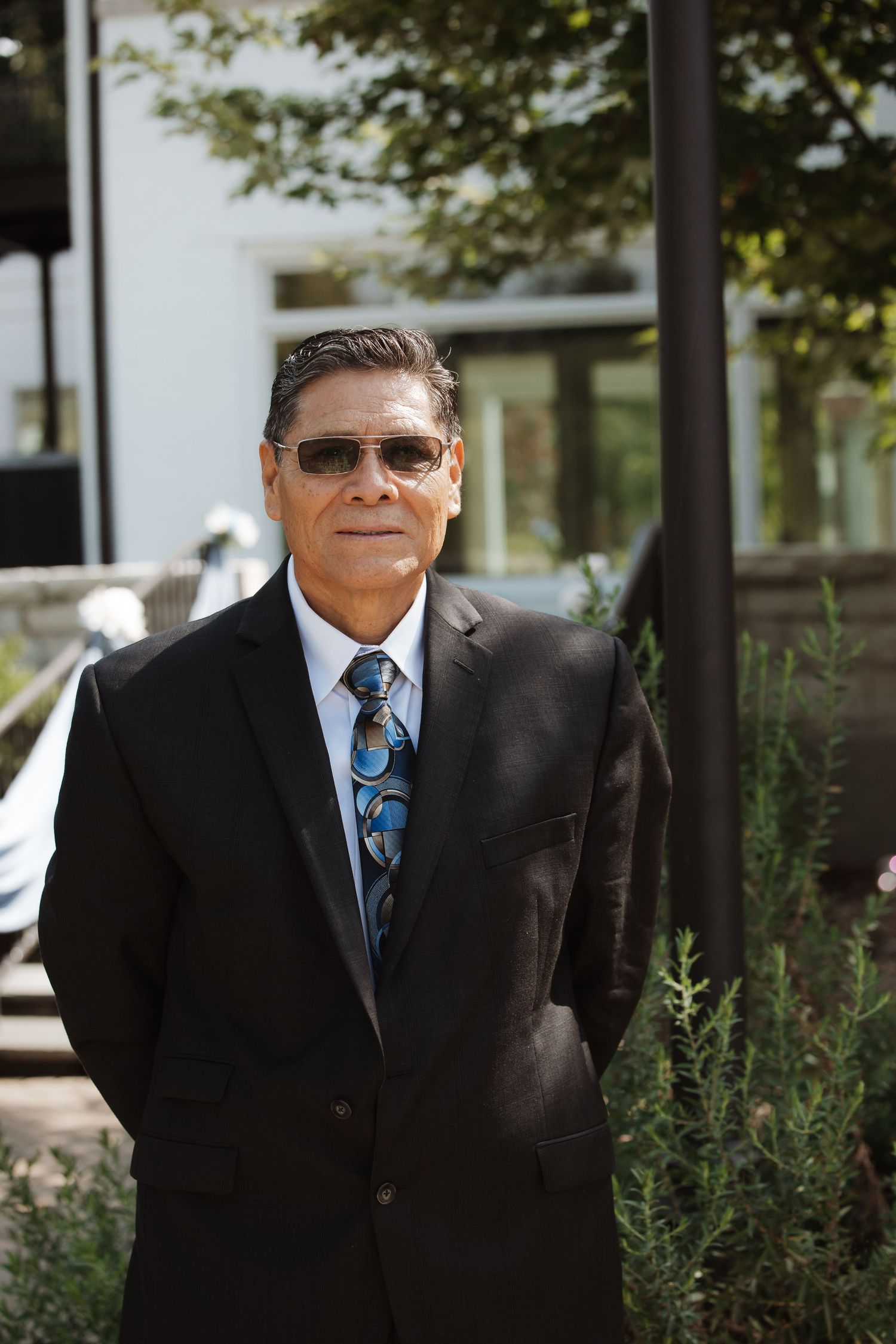 Elegant older man in black suit and patterned tie wearing sunglasses outdoors at wedding.