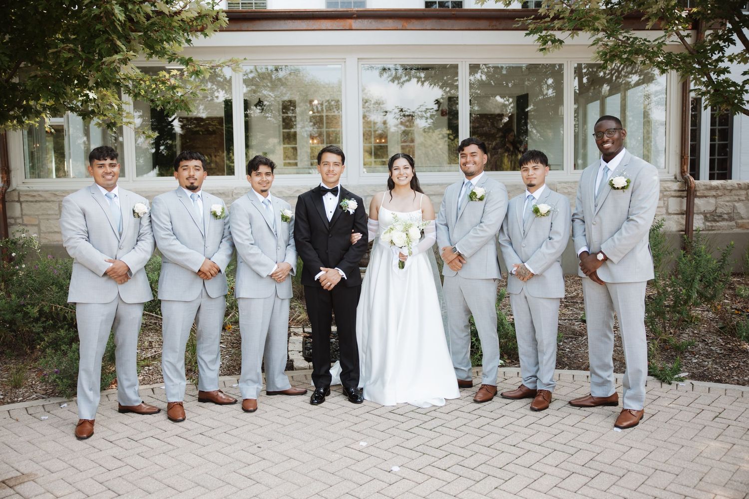 Groom in black tuxedo stands with bride and seven groomsmen in light gray suits outside venue.