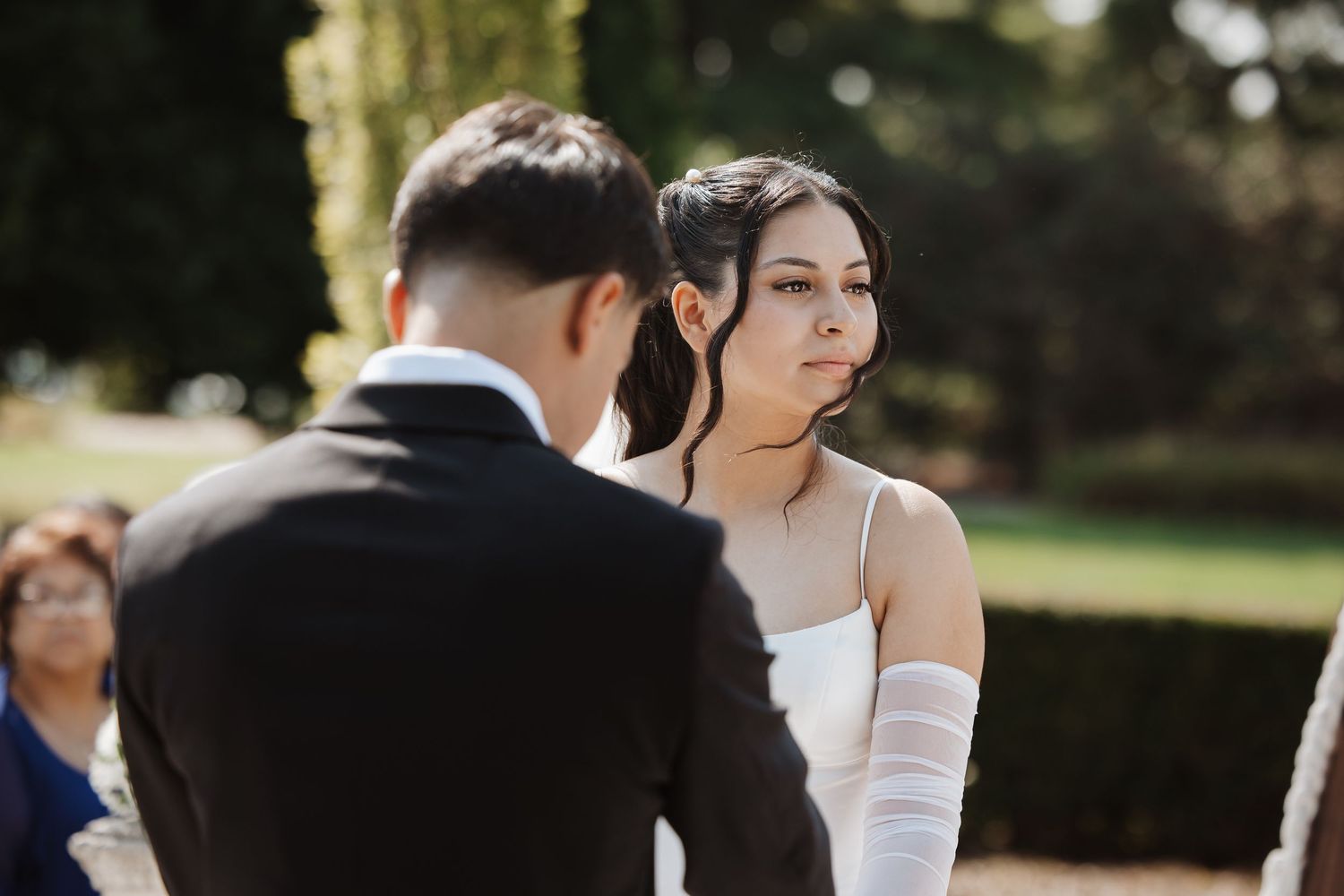 A bride in a white gown with white gloves looks lovingly at the groom during an outdoor ceremony.