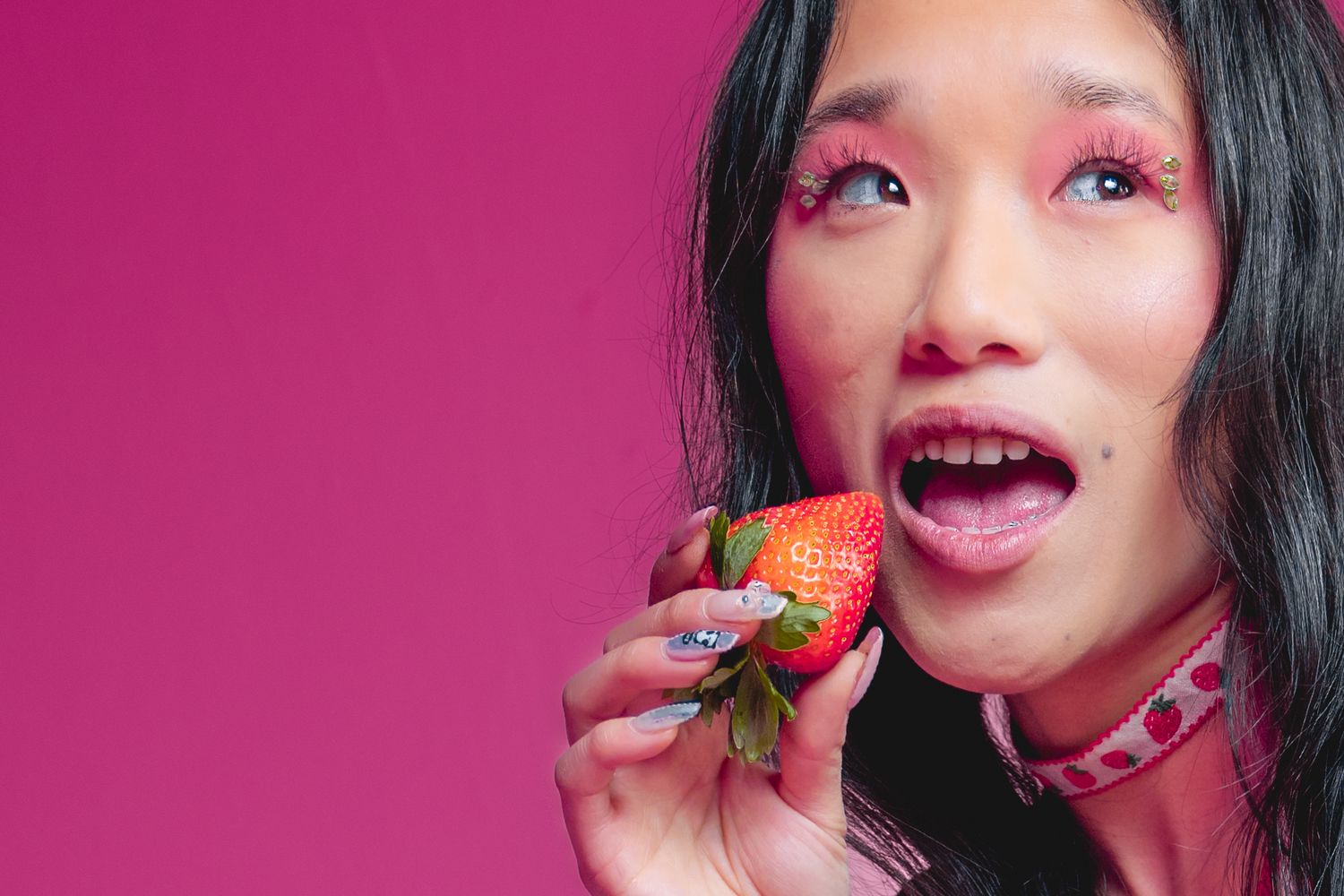 Asian woman with pink eye makeup holds large strawberry near open mouth on pink background.