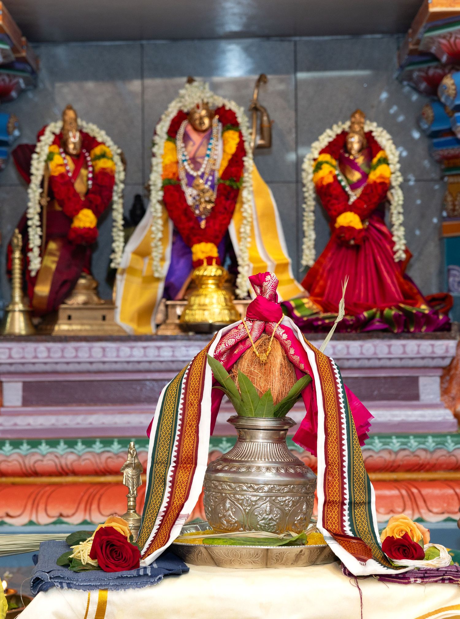 Hindu temple altar with decorated brass kalash and three deity idols adorned with flowers and garlands.