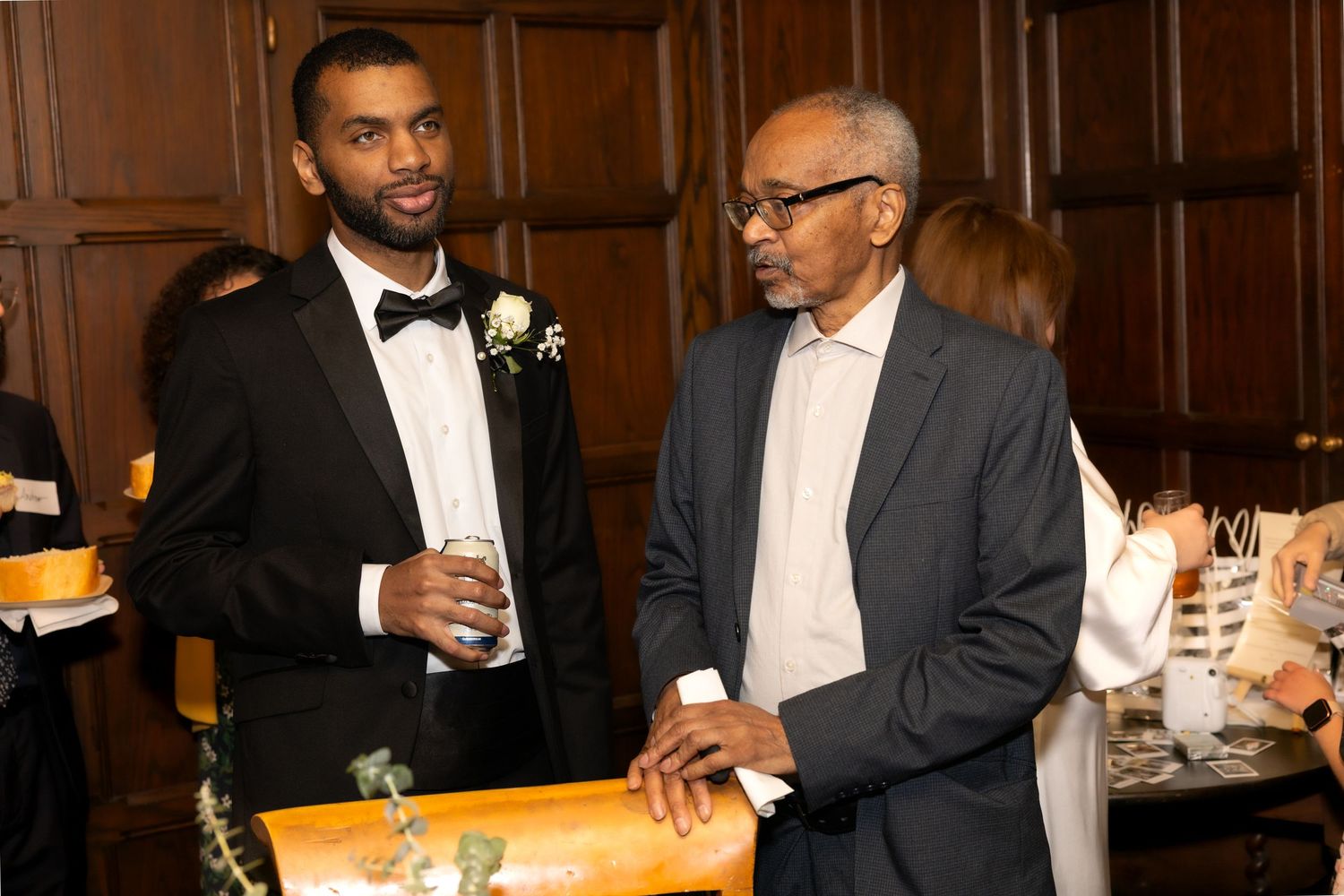 Groom in tuxedo with boutonniere converses with older gentleman at wedding reception.