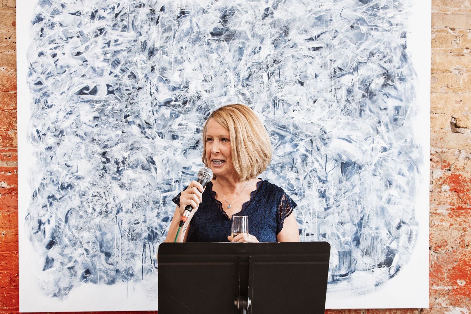 Blonde woman in navy dress speaks at microphone podium before large blue abstract painting.