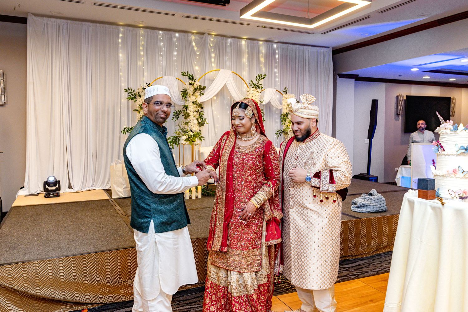 A man in teal vest congratulates a South Asian bride and groom in traditional wedding attire at reception.