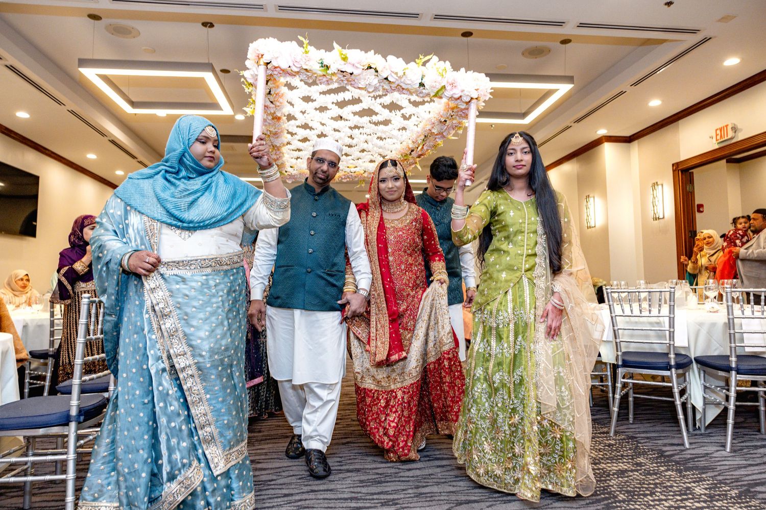 South Asian bride in red lehenga walks under flower canopy with family during traditional wedding entry.