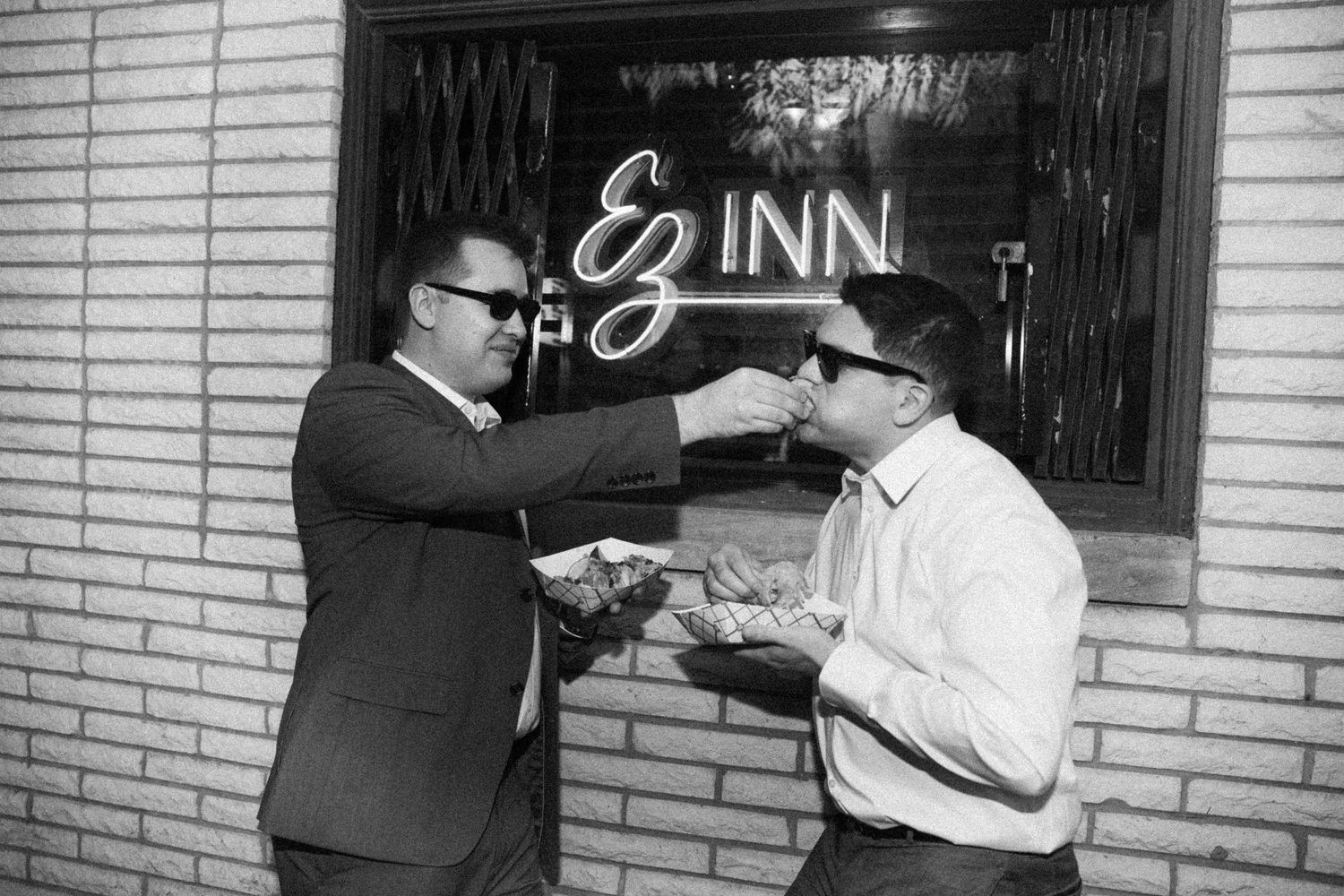 Black and white photo of two men in suits sharing food outside EZ Inn entrance.