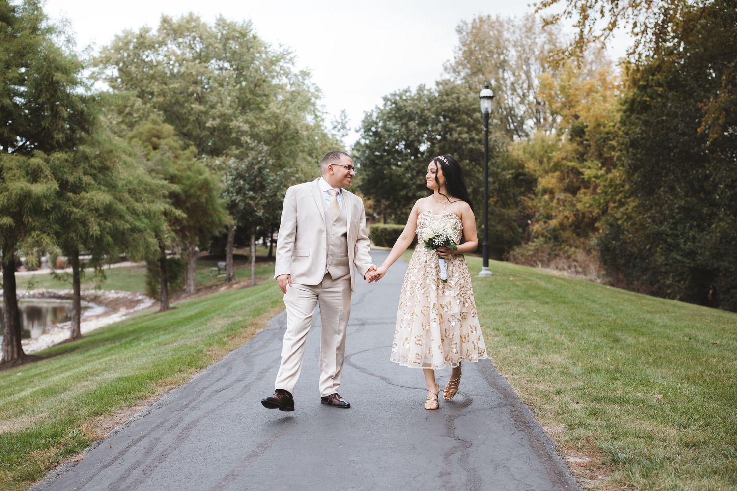 Bride in floral dress and groom in tan suit holding hands while walking on a tree-lined path outdoors.