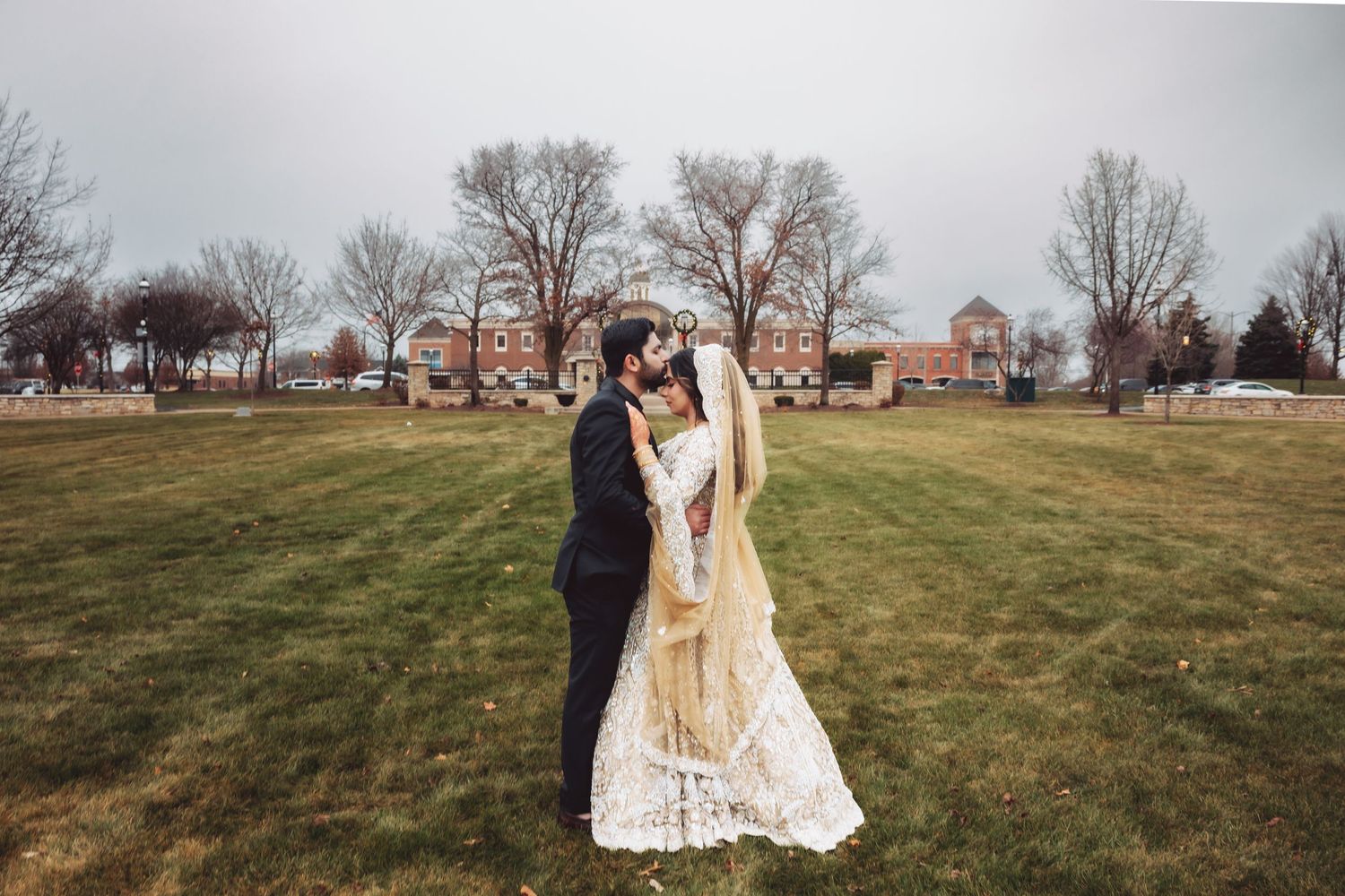 Pakistani bride and groom embrace tenderly on green lawn with brick building backdrop in winter setting.