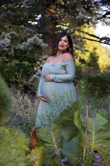 Pregnant woman in sage green off-shoulder maternity gown posing among lush garden greenery and wildflowers.
