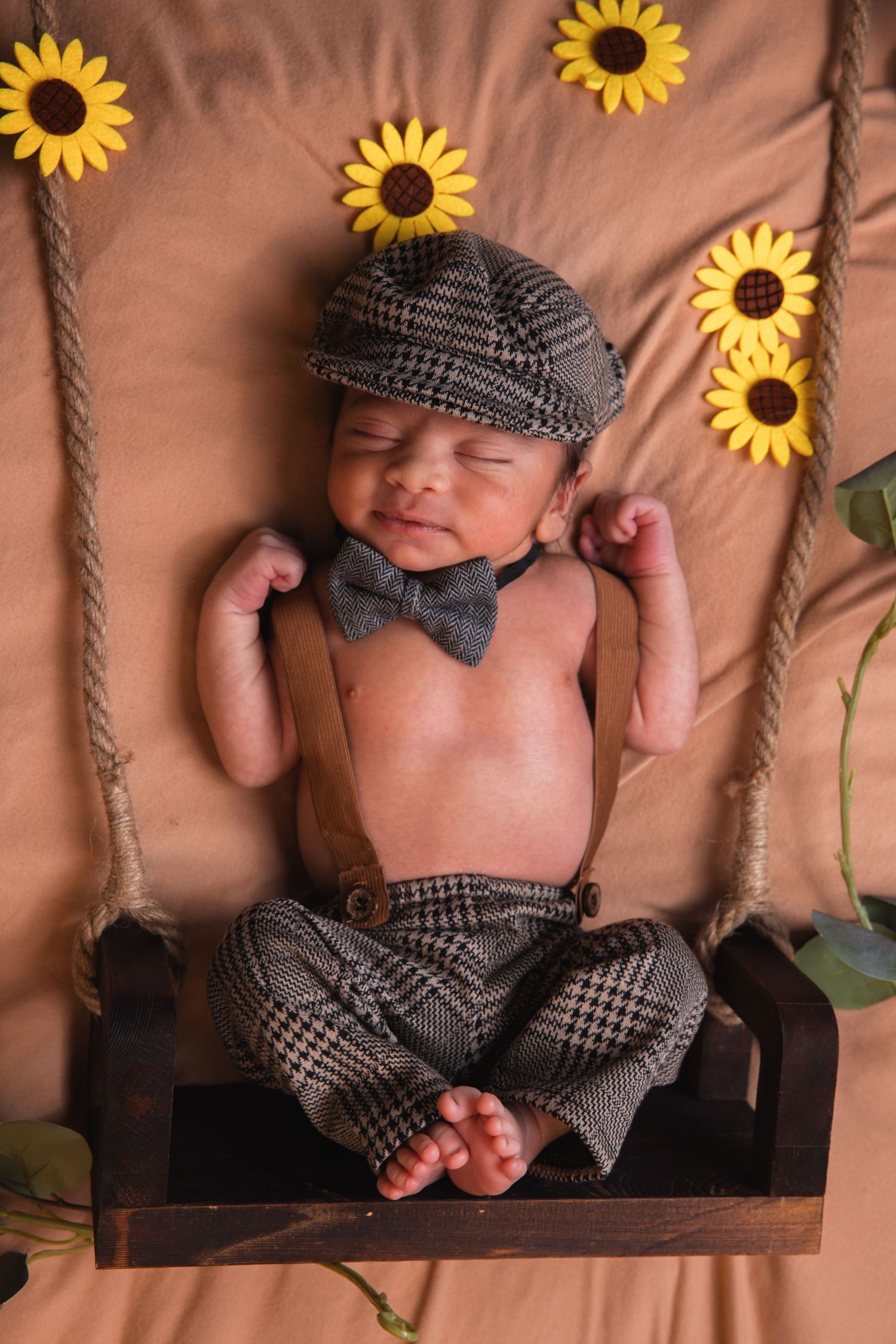 Newborn baby in vintage tweed cap and pants sits on wooden swing with sunflower decorations.