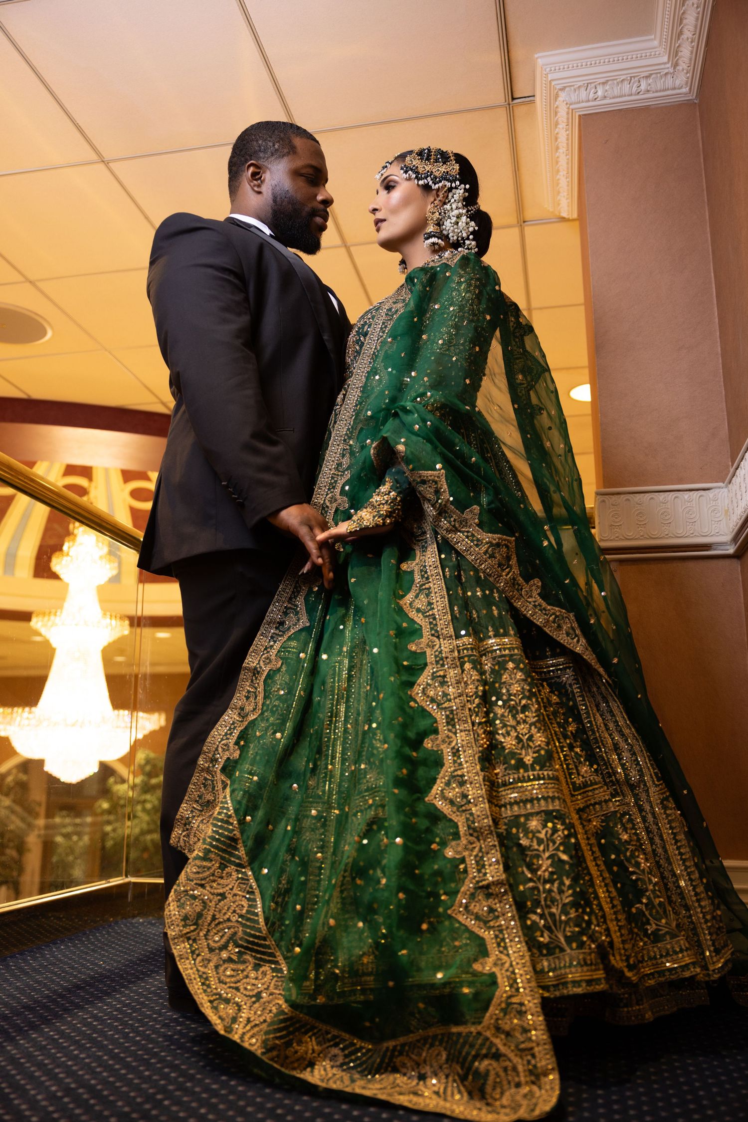 Bride in green embroidered lehenga and groom in black tuxedo share a romantic pose indoors.
