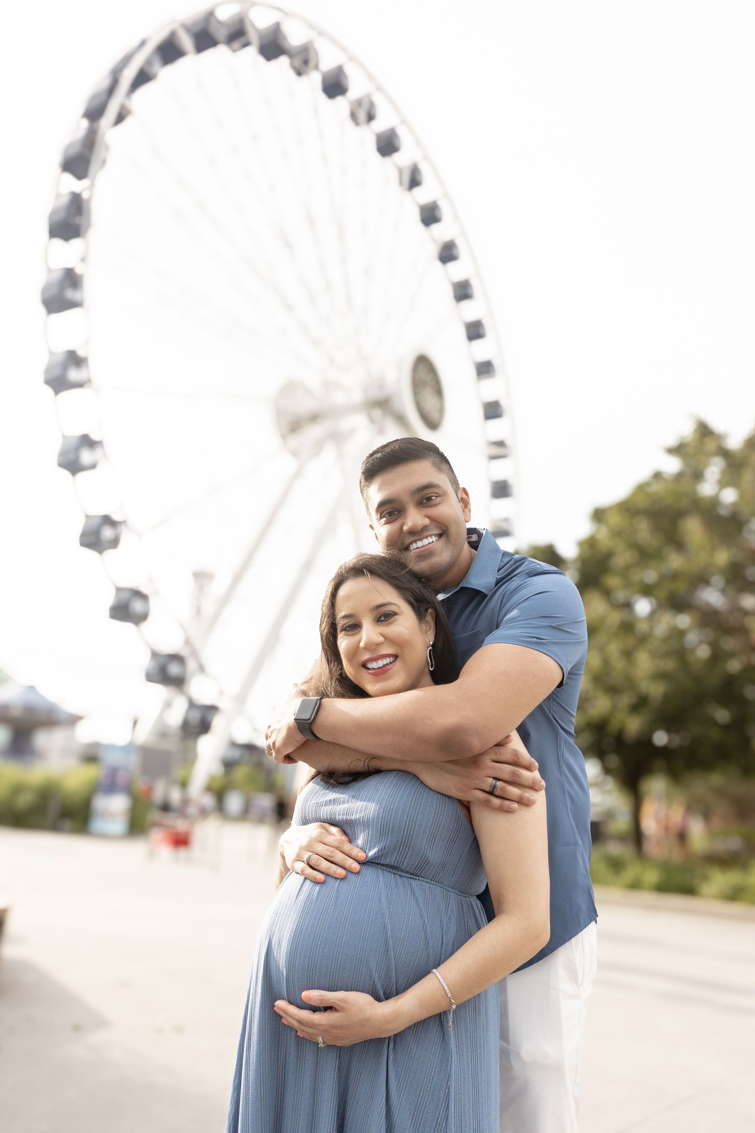 Smiling couple embracing during outdoor maternity photo near Ferris wheel.