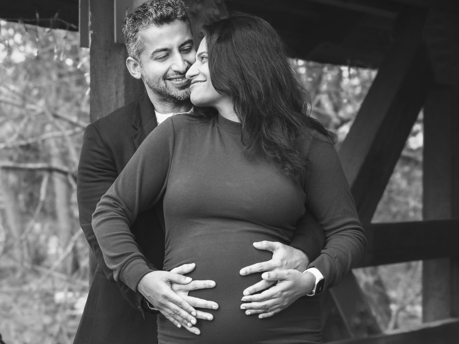 Black and white maternity portrait of man kissing pregnant womans cheek outdoors.