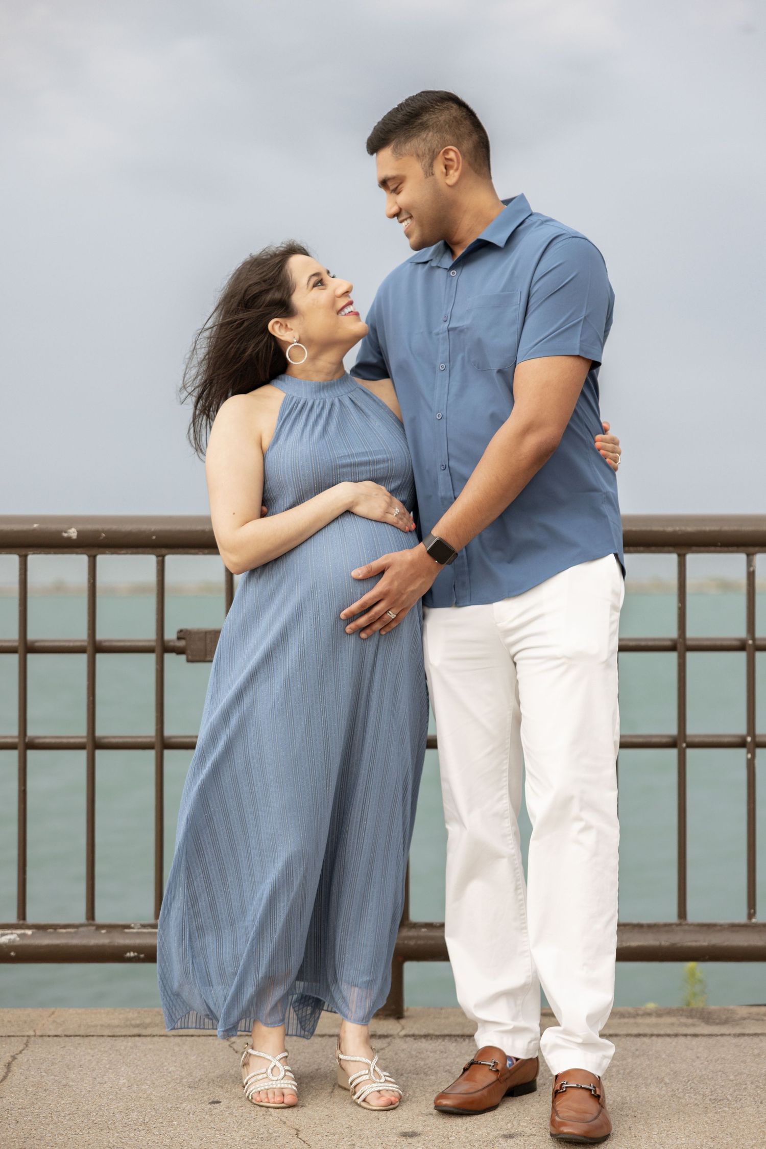 Smiling pregnant couple in matching blue outfits embracing on a waterfront pier, gazing lovingly at each other.