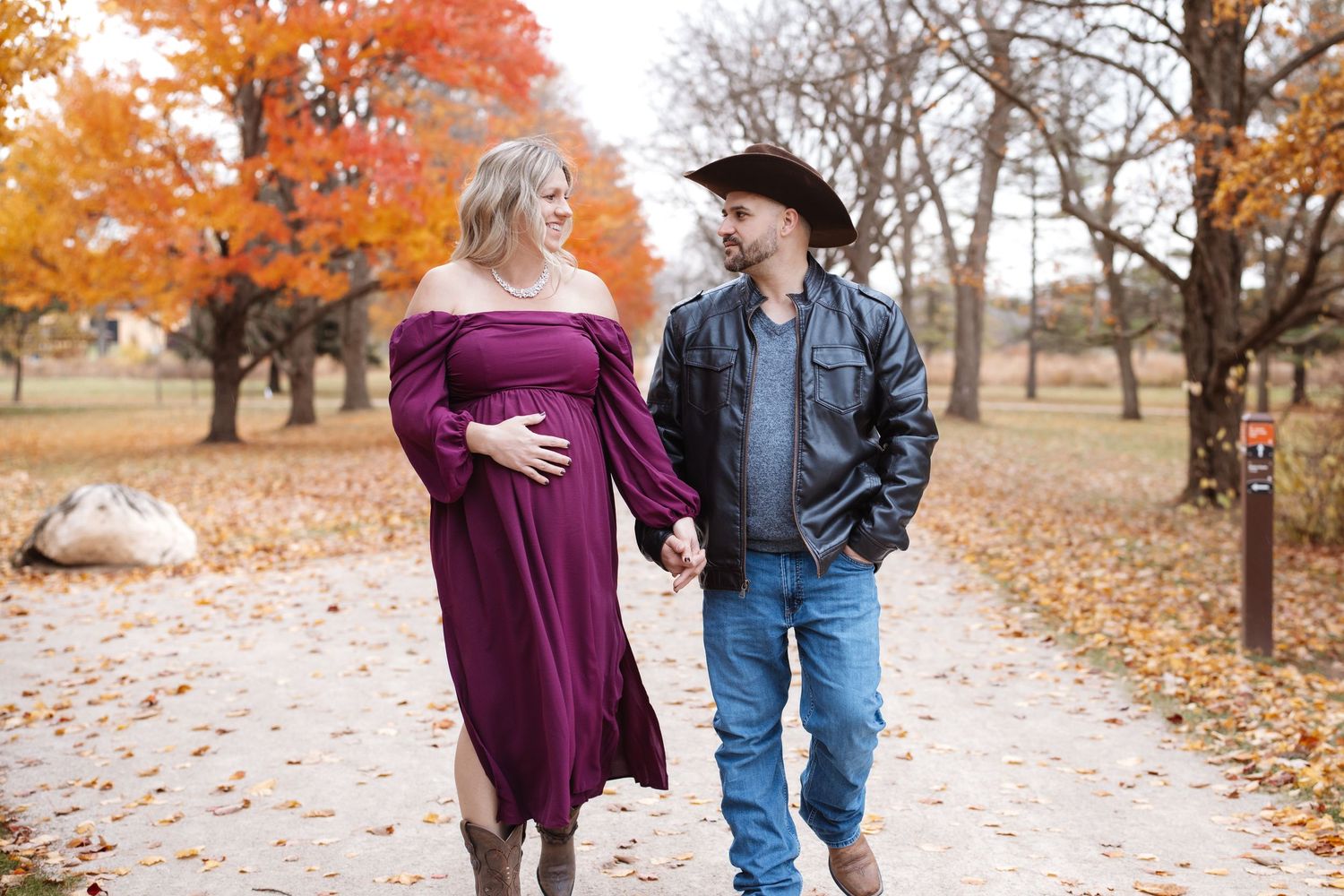 Expecting couple holding hands on leaf-lined autumn path with colorful fall trees behind them.