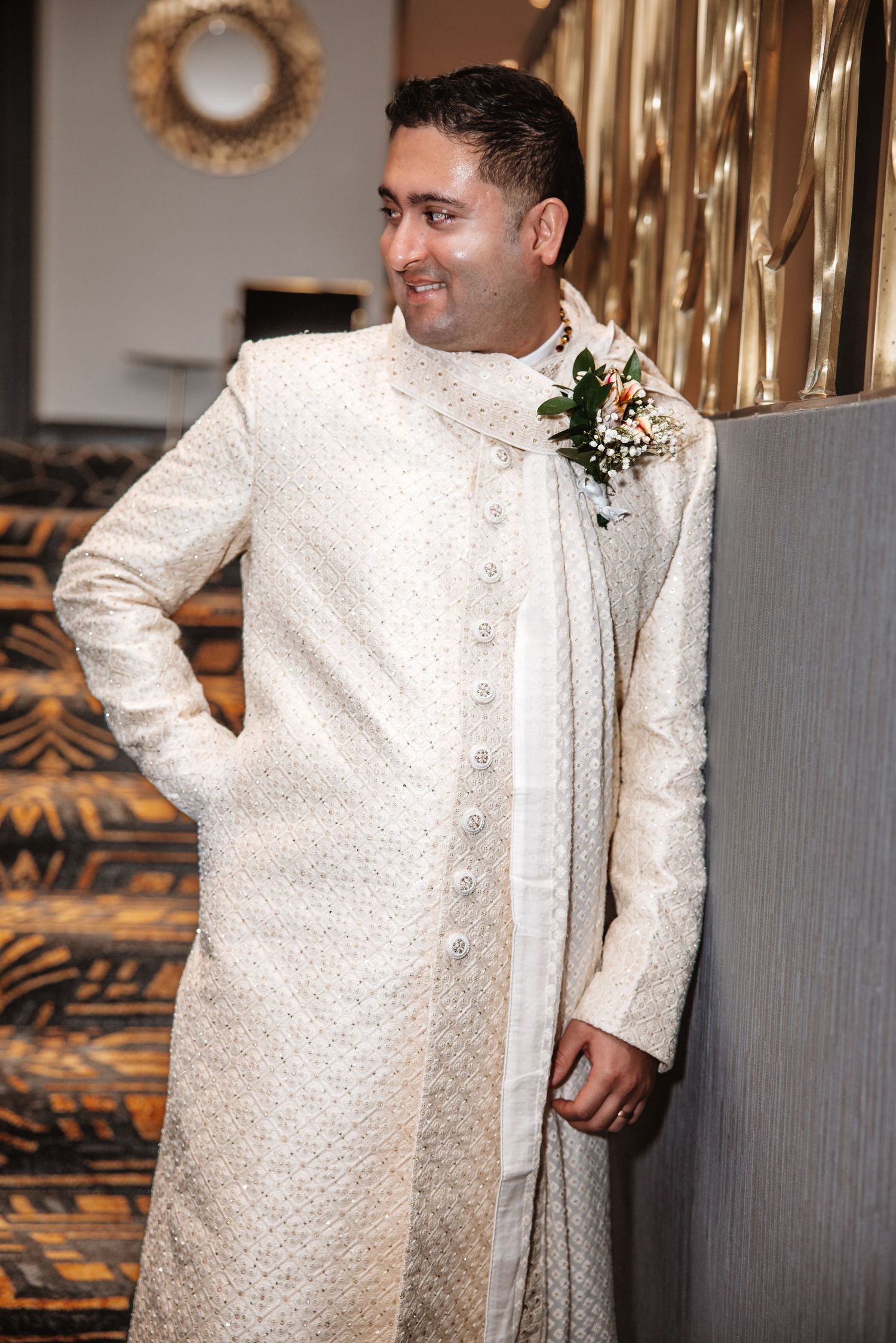 Dapper groom in cream sherwani with boutonniere smiles while leaning against golden pillar.