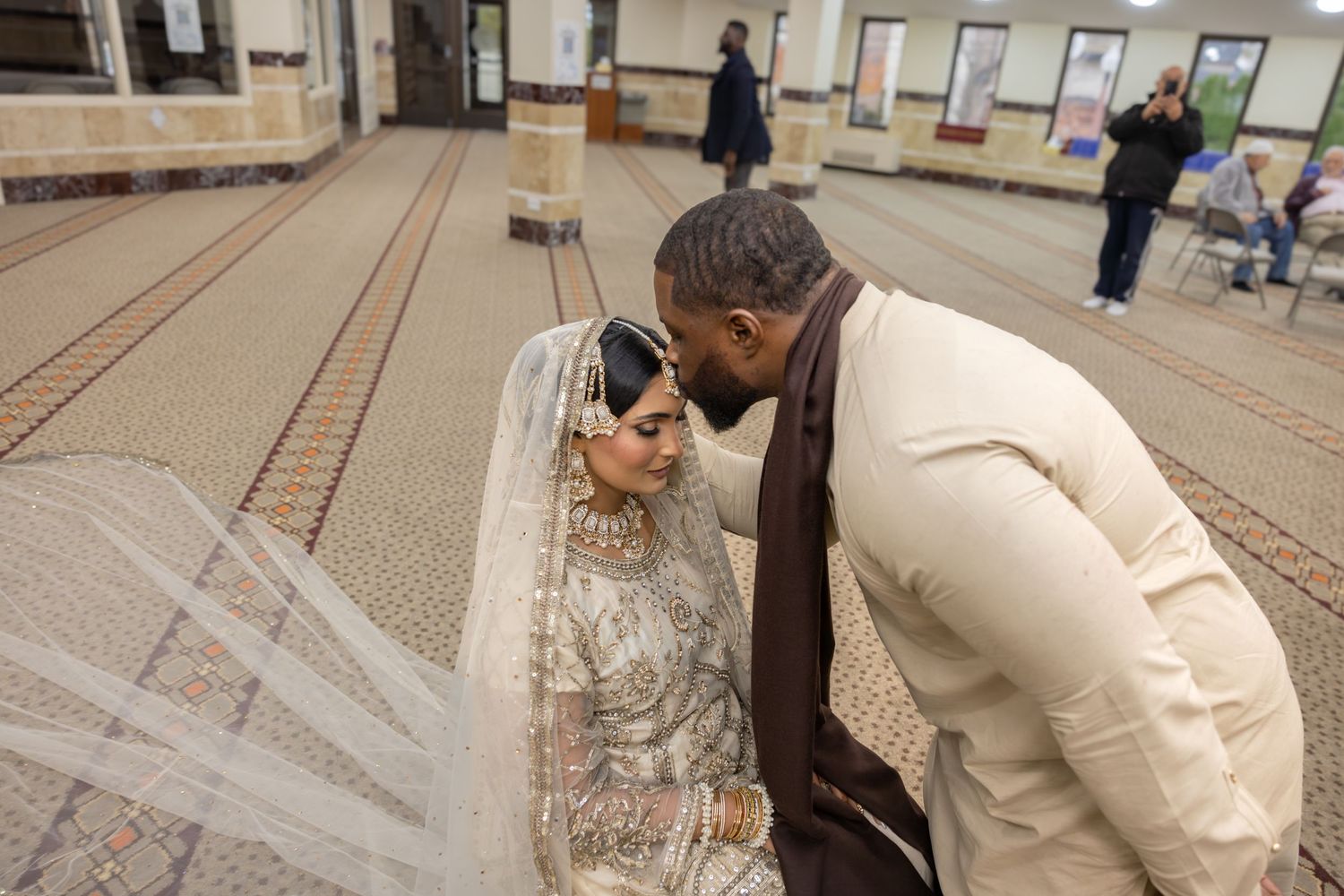 Groom tenderly leaning toward veiled bride in ornate white lehenga during intimate nikah ceremony in mosque.