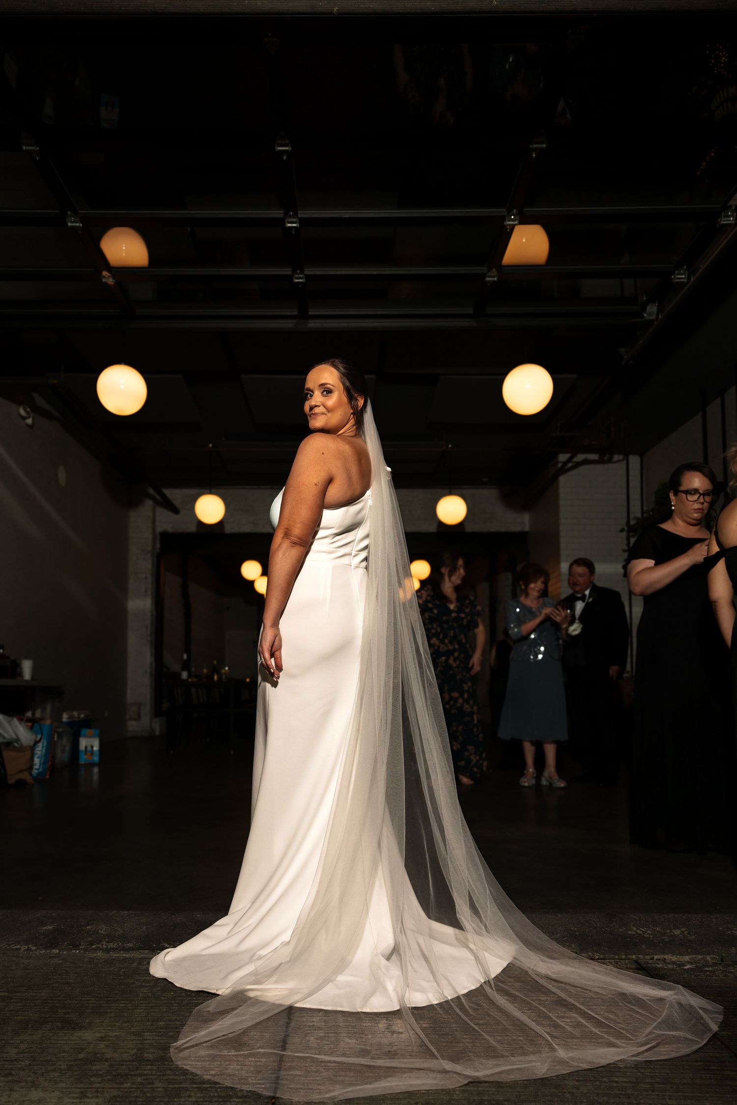 Beautiful bride in fitted white gown with long flowing veil looks back over shoulder in dramatic industrial setting.