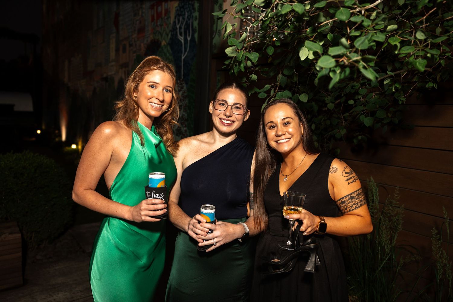 Three women in elegant green and black dresses holding drinks at an outdoor evening wedding reception.