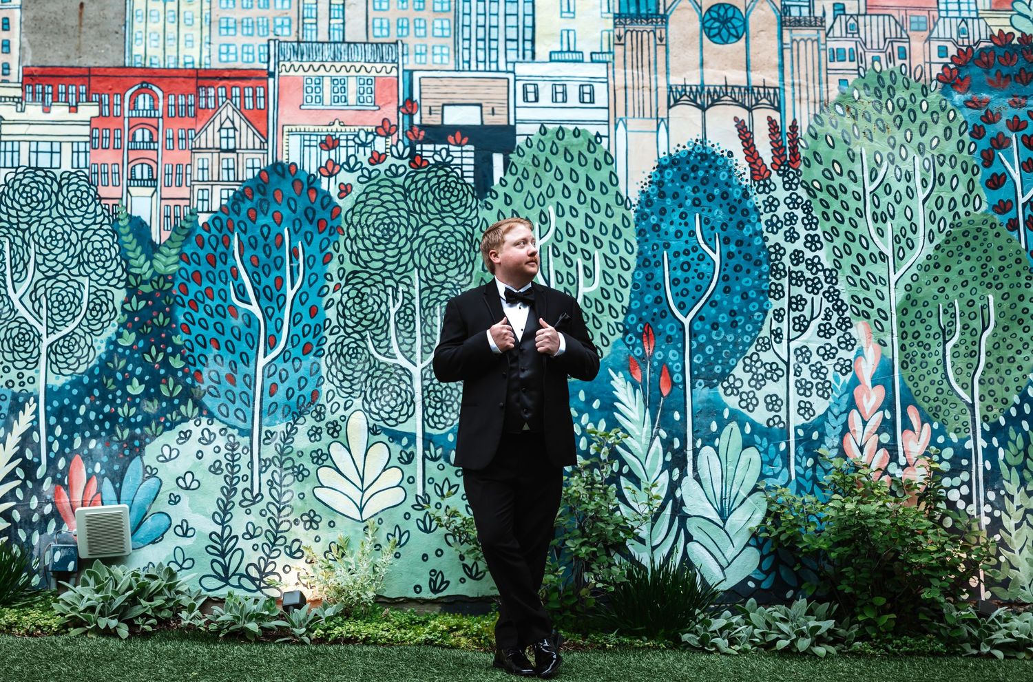 Groom in black tuxedo stands smiling before a large colorful cityscape and garden mural backdrop.