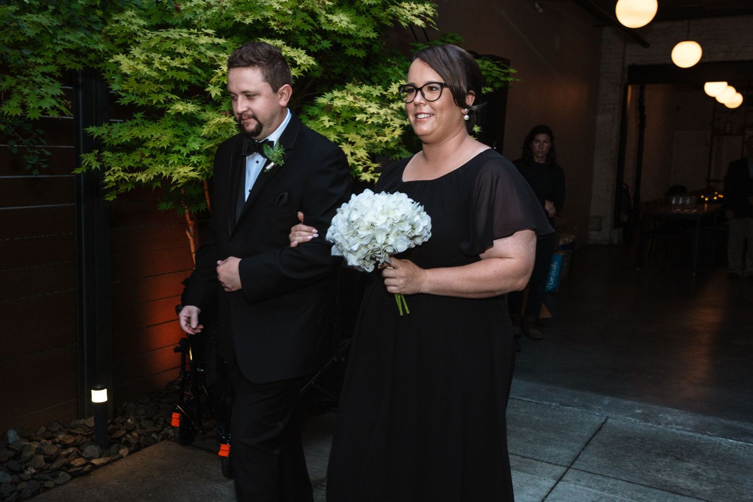 A man in a tuxedo escorts a woman in a black gown holding white hydrangeas at a wedding ceremony.