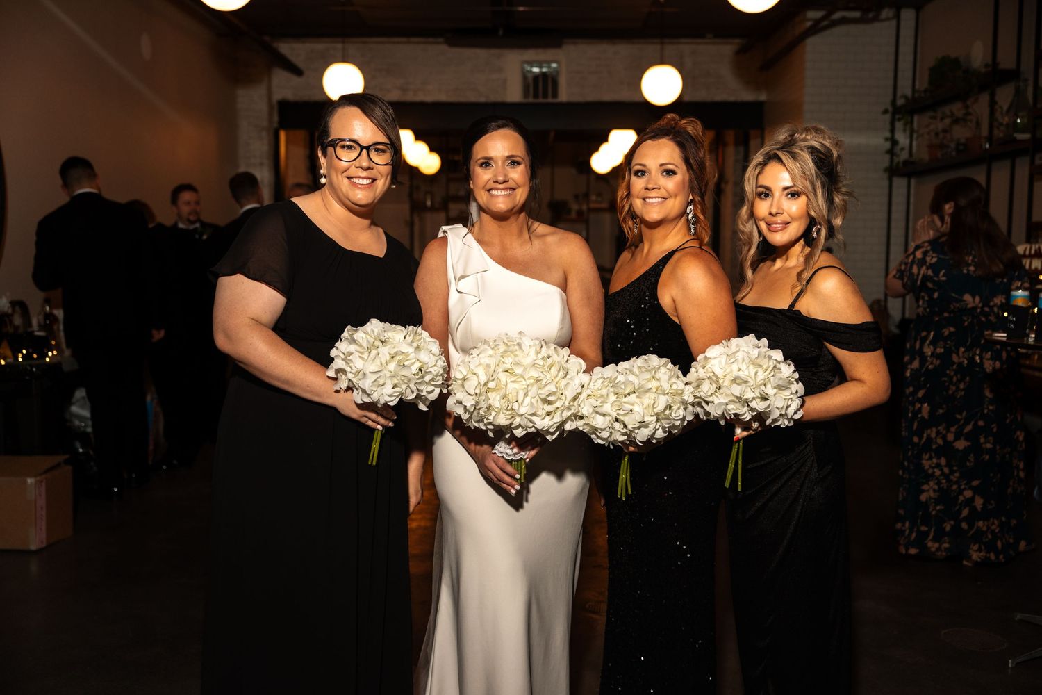 Bride in one-shoulder white gown posing with three bridesmaids in black dresses holding white bouquets.