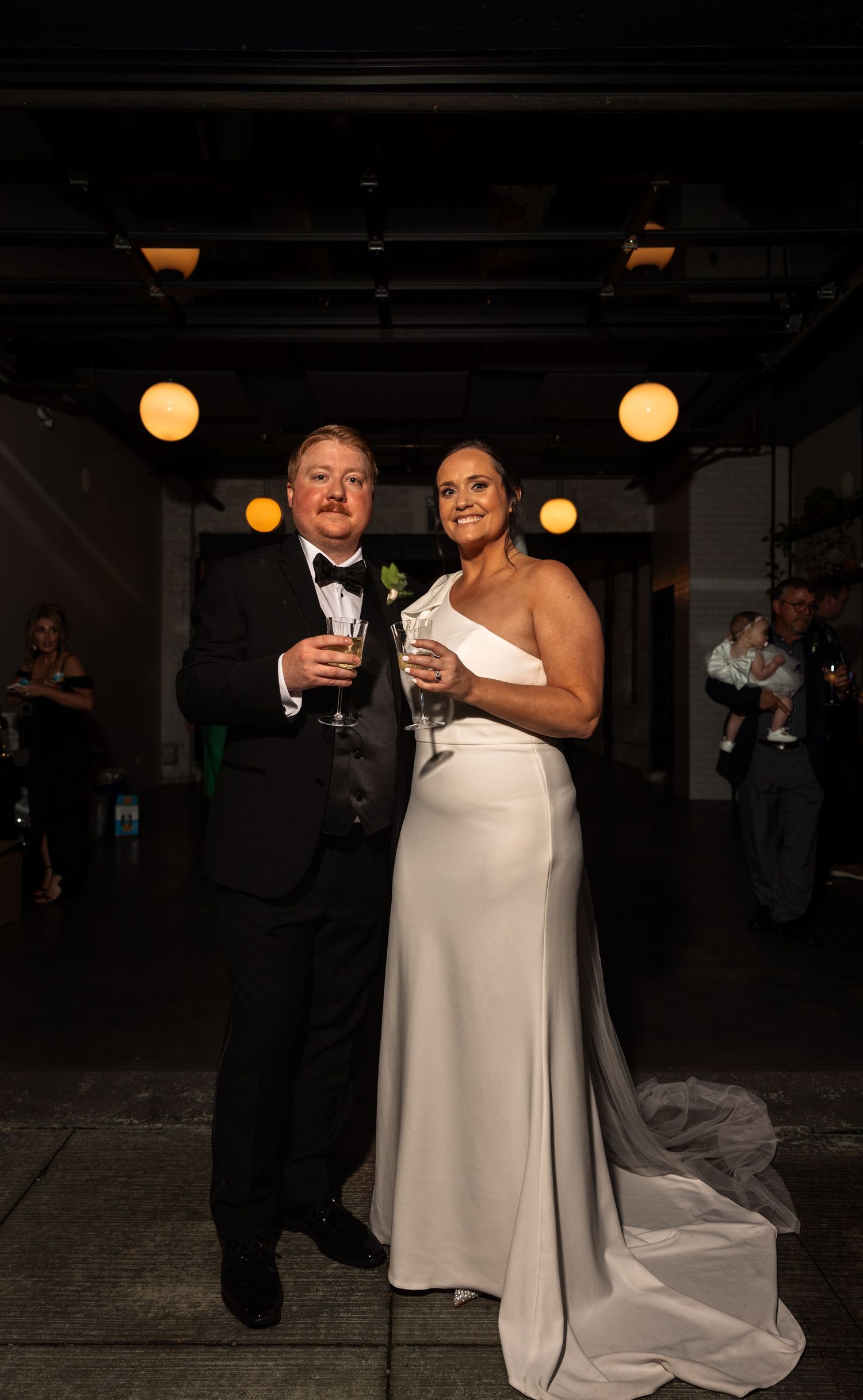 Bride in white gown and groom in black tuxedo pose with champagne at elegant wedding reception.