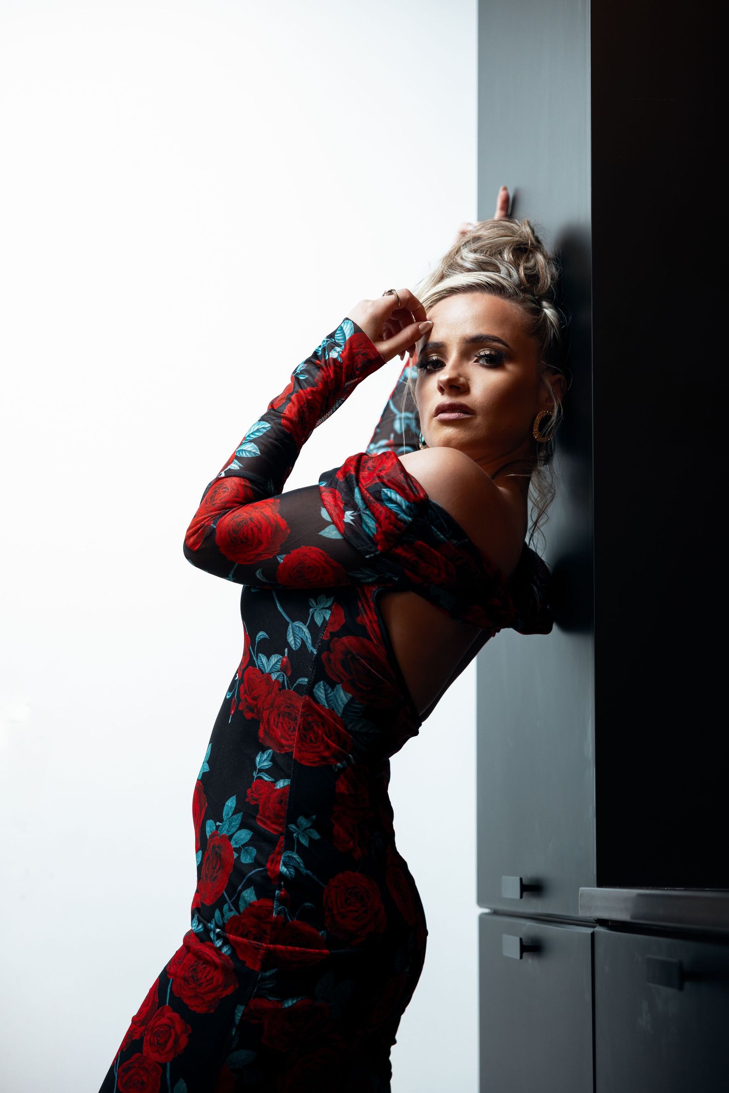 Elegant blonde woman in backless red floral maxi dress poses leaning against dark architectural wall.