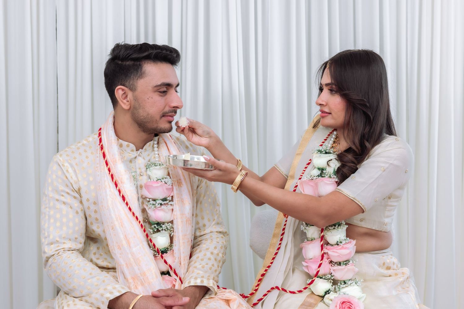 Indian bride in white lehenga playfully feeds groom during traditional wedding ceremony while both wear floral garlands.