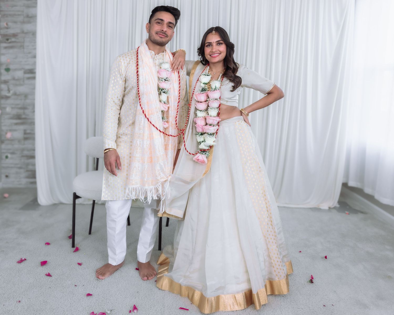 Indian bride and groom in matching white and gold traditional wedding attire adorned with pink and white floral garlands.