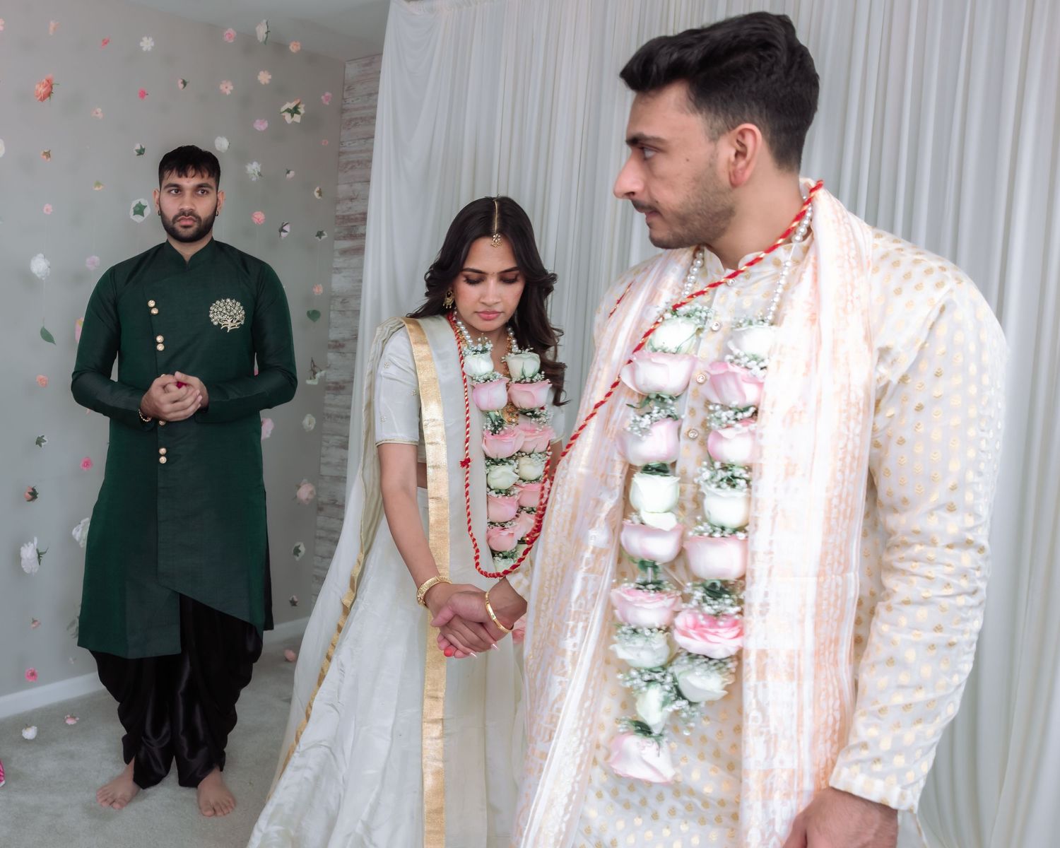 Indian bride and groom in traditional wedding attire with guest in green kurta.
