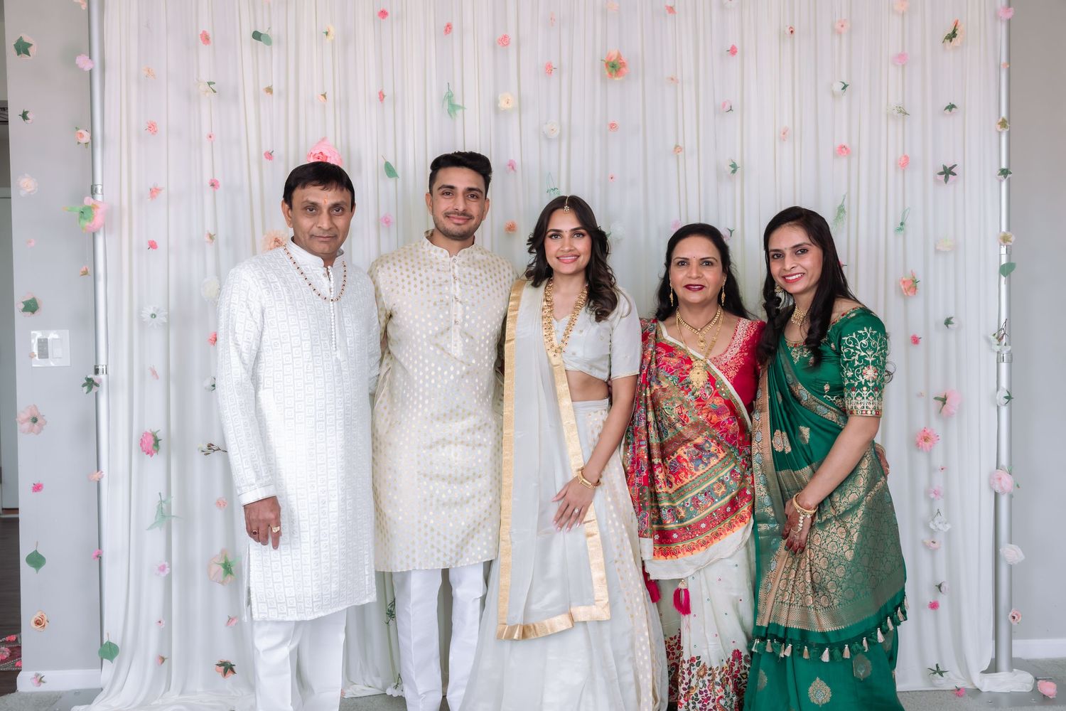 Five people in traditional Indian attire including white lehenga and colorful sarees pose at a wedding celebration.