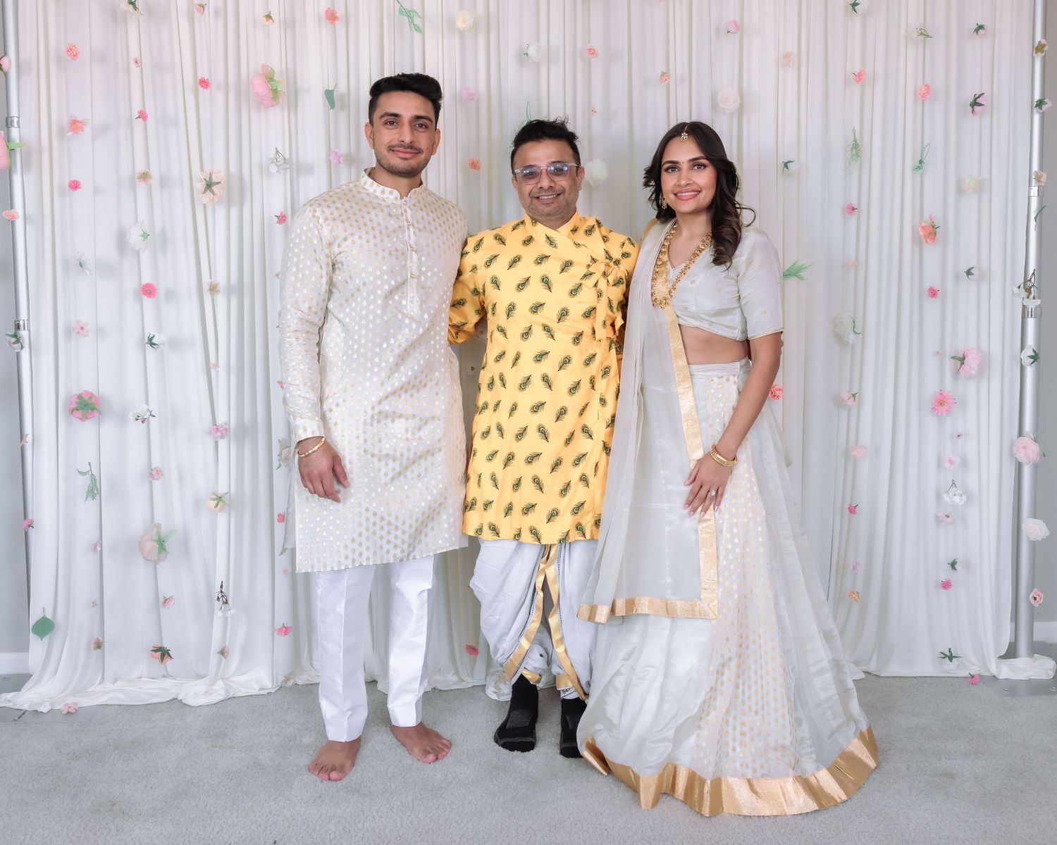 Three young adults in traditional Indian attire including white and yellow outfits pose at festive event.