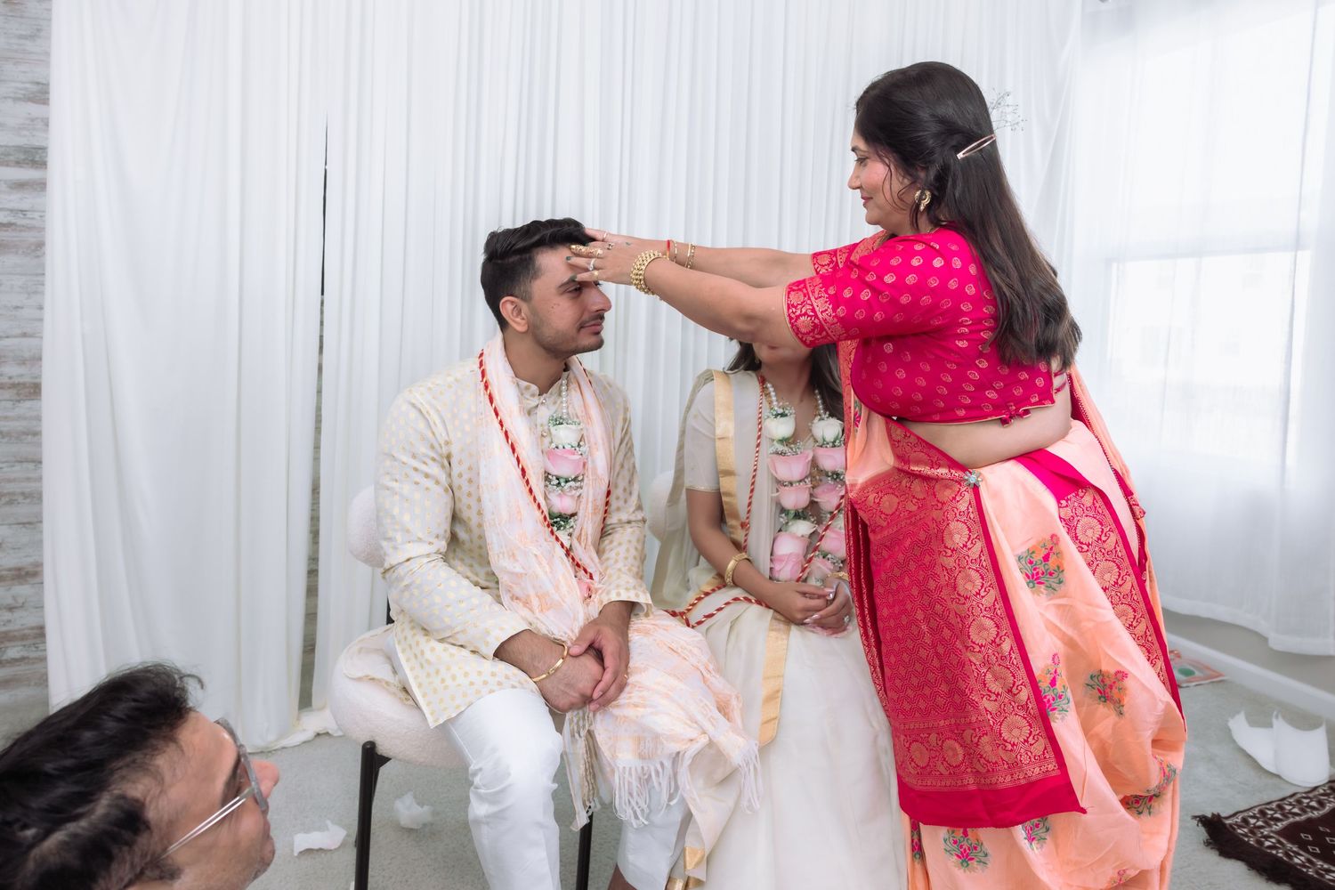 A woman applies tilak to a groom during a traditional Indian wedding ceremony indoors.