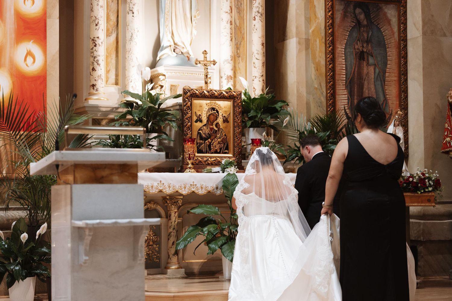 Bride in white gown and veil praying before ornate church altar with religious icons and floral arrangements.