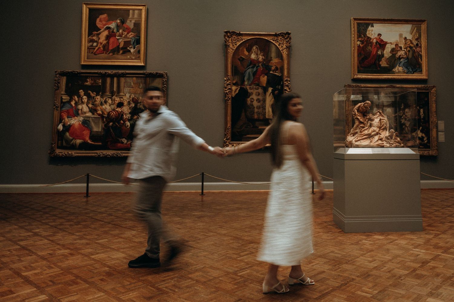Couple holds hands dancing through sunlit art museum gallery surrounded by Renaissance paintings.