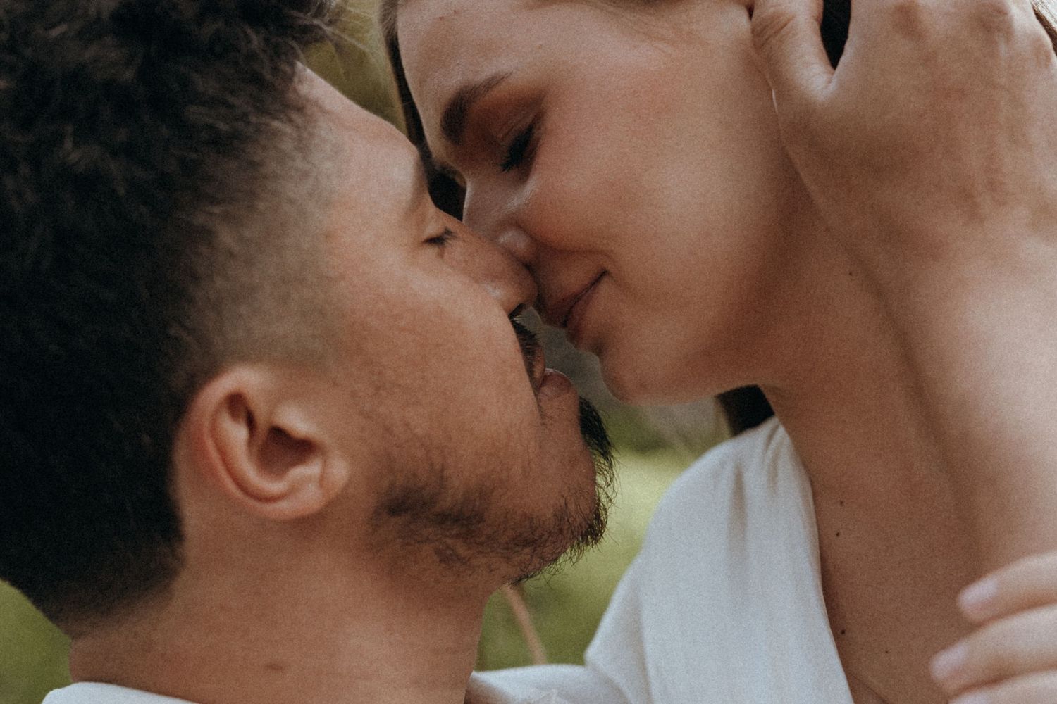 A couple shares a tender romantic kiss outdoors, captured in an intimate close-up portrait.