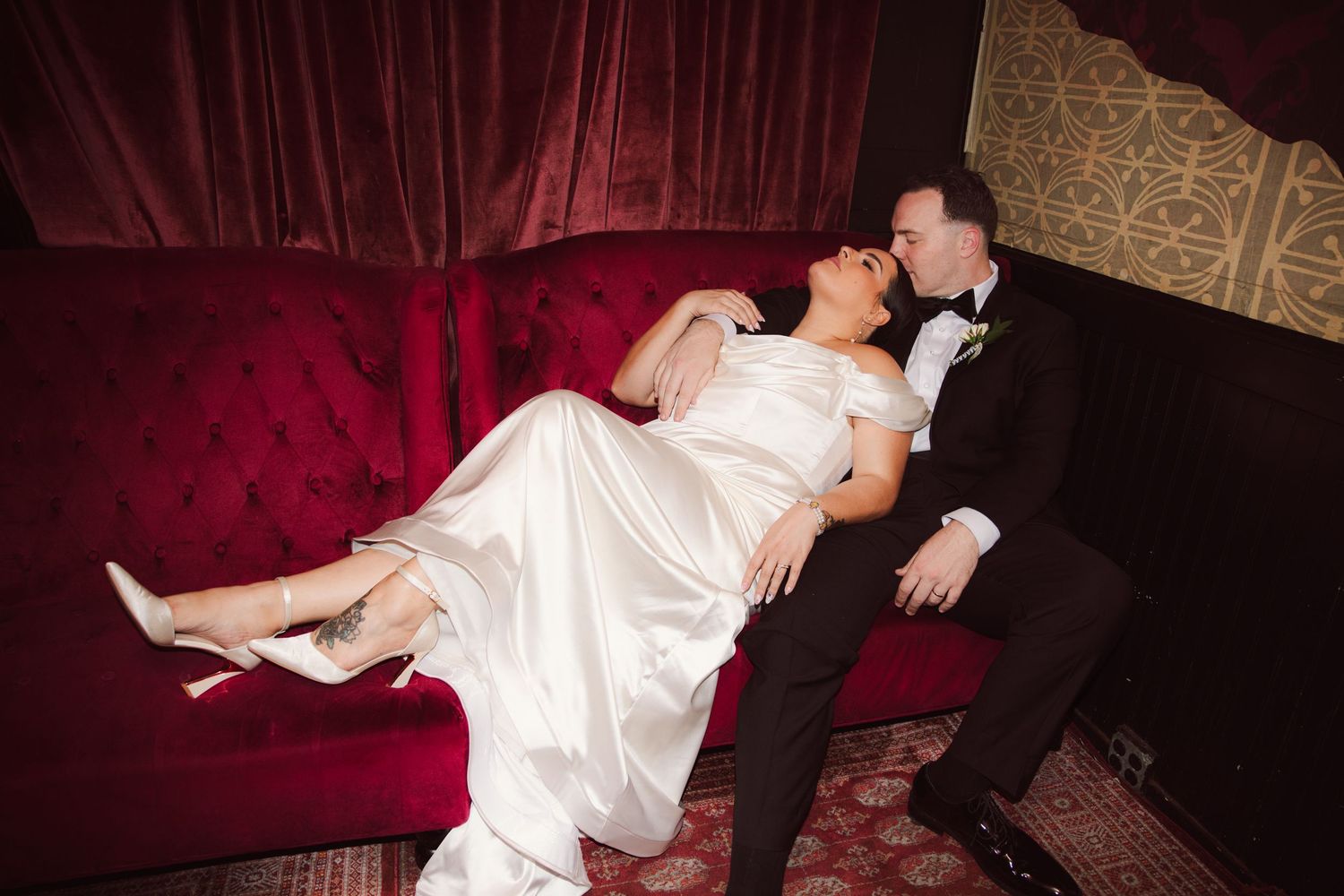 Bride and groom sharing a romantic moment on a red velvet sofa at their wedding reception.