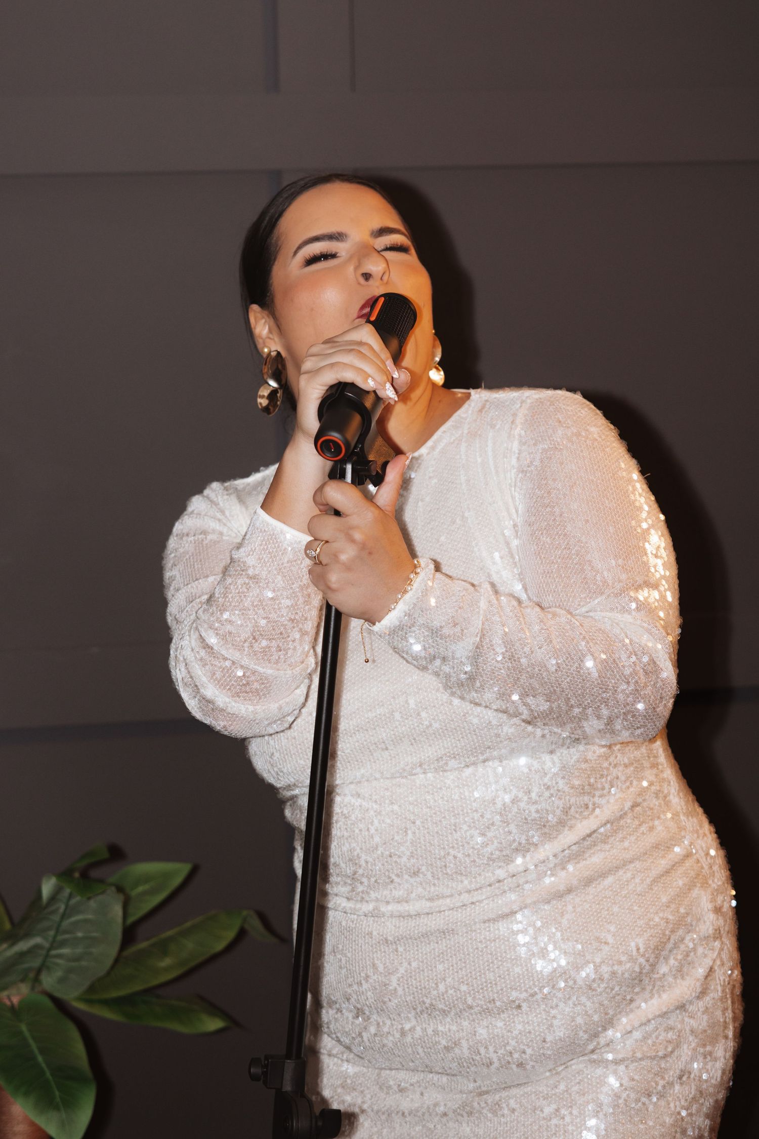Woman in white sequin dress singing passionately into a microphone at an indoor event.