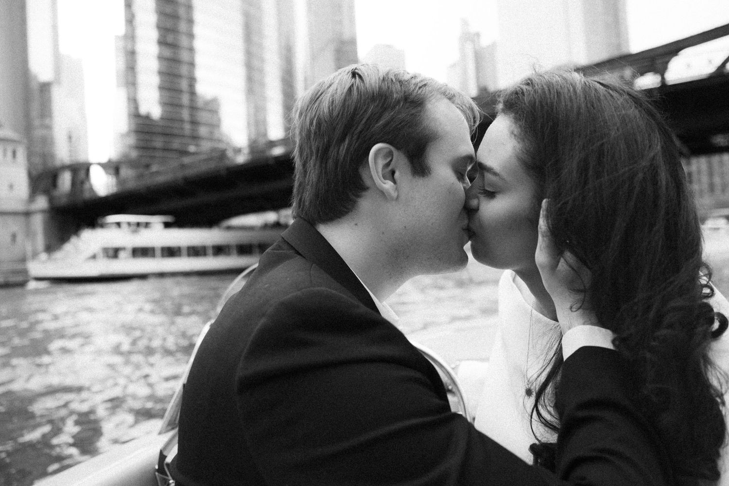Black and white photo of a couple kissing on a boat with a city skyline and bridge backdrop.