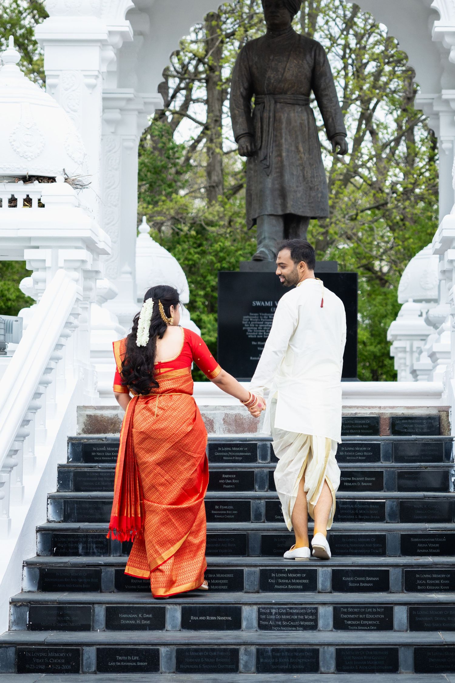 Indian wedding couple ascending decorated white temple stairs hand in hand, bride in red saree and groom in white.