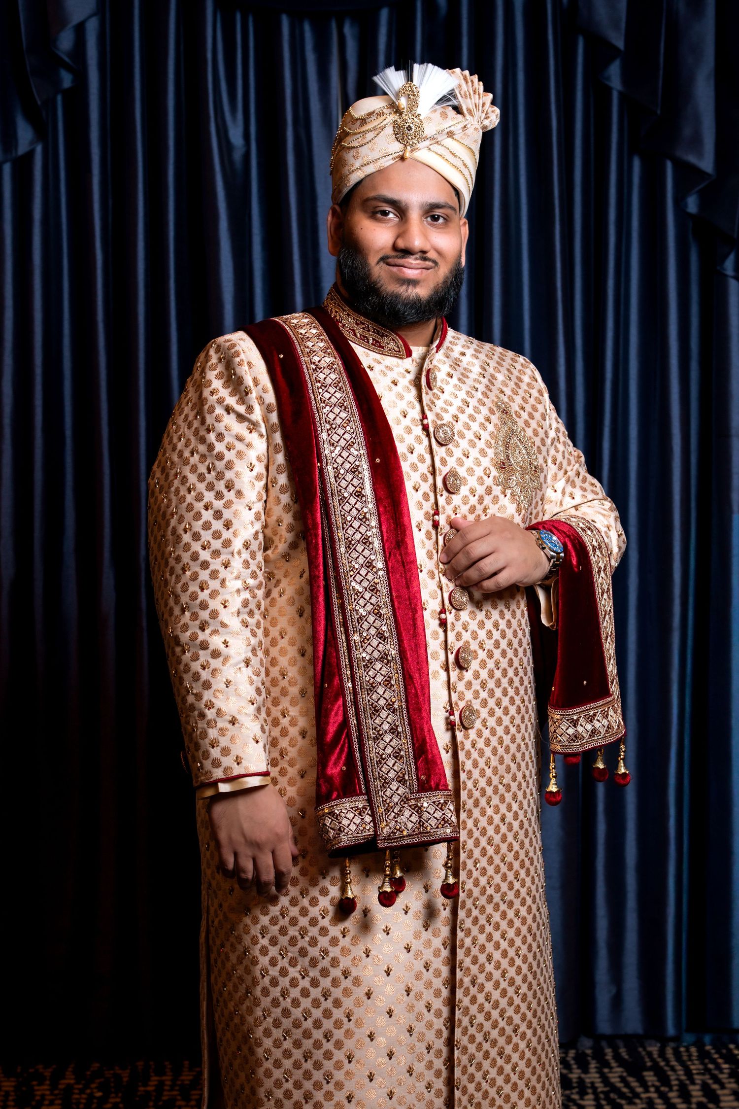 South Asian groom in embroidered gold sherwani with red dupatta and decorative turban against blue curtains.