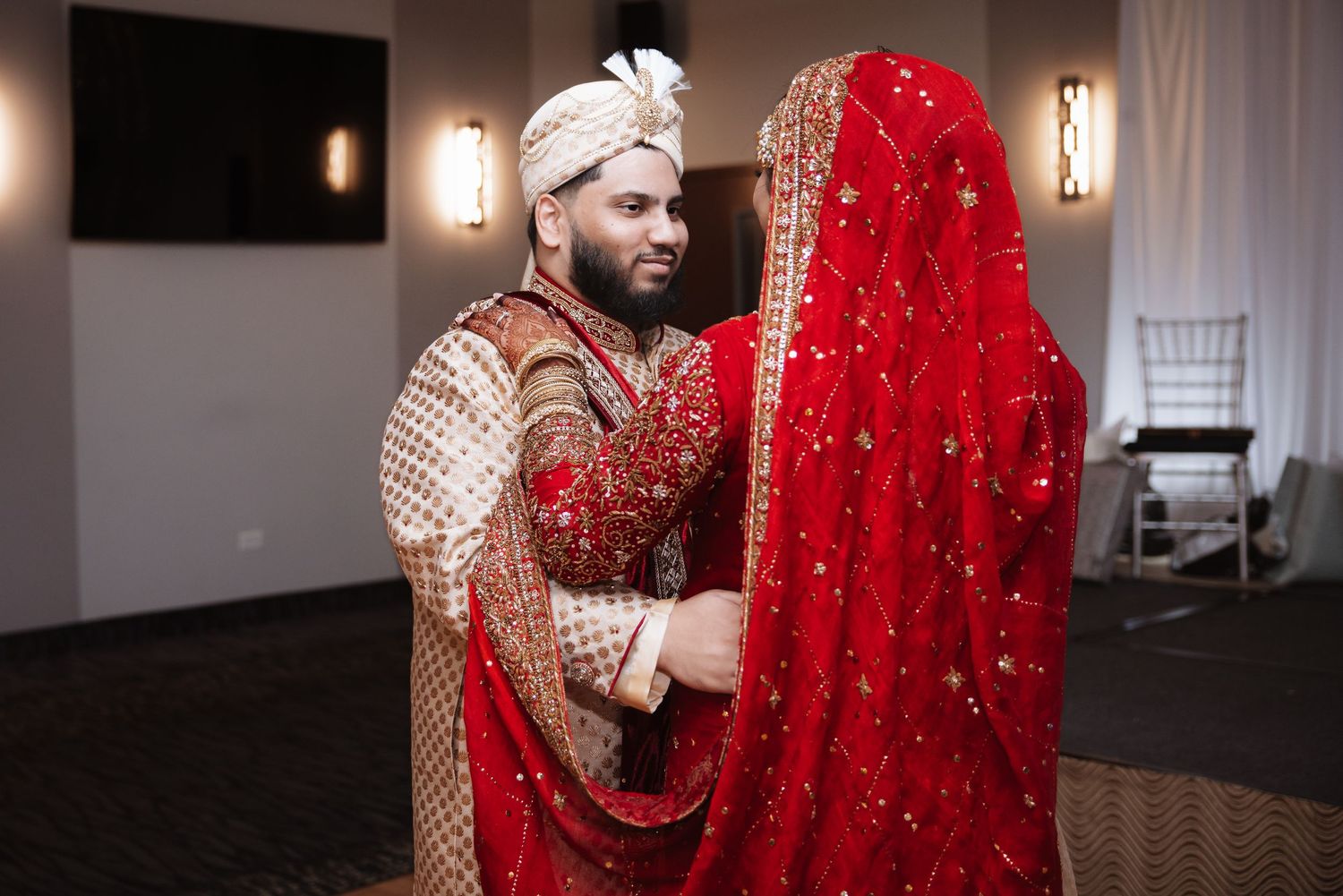 Groom in traditional South Asian sherwani tenderly holds bride in ornate red bridal veil.