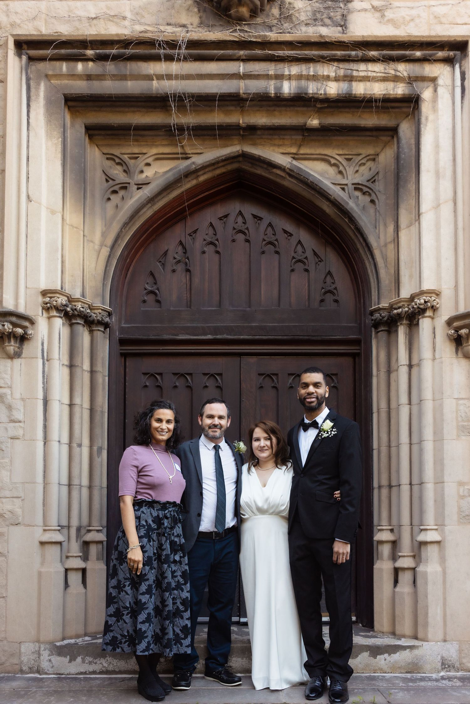 Wedding couple and two guests pose before ornate Gothic stone church doorway.