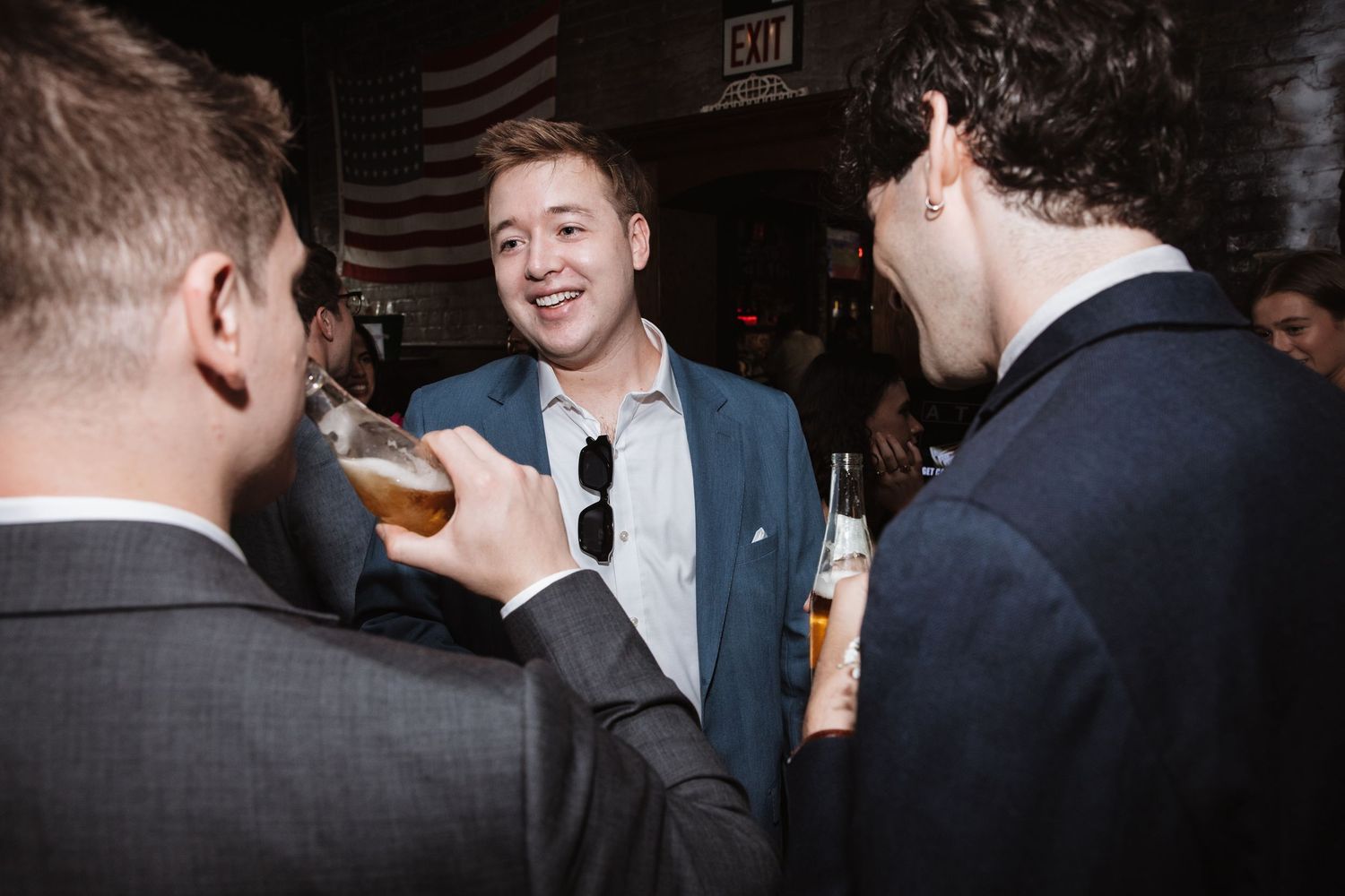 Three men in suits smile and chat while holding beers at a wedding reception.