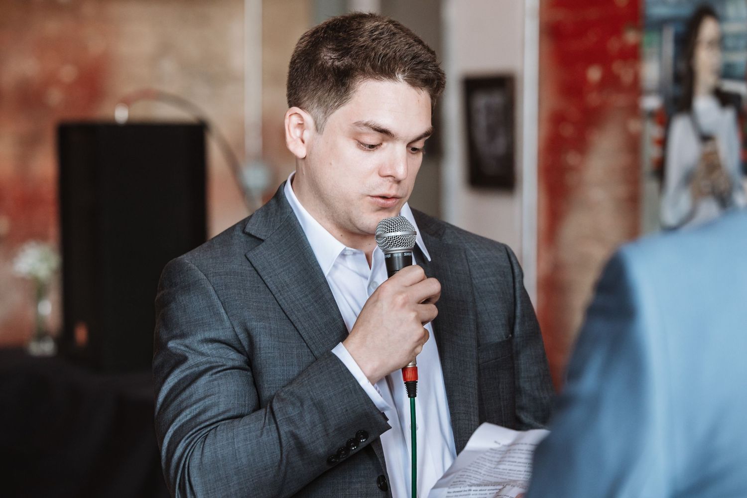 Young man in a gray suit speaking into a microphone while holding papers at a formal event.