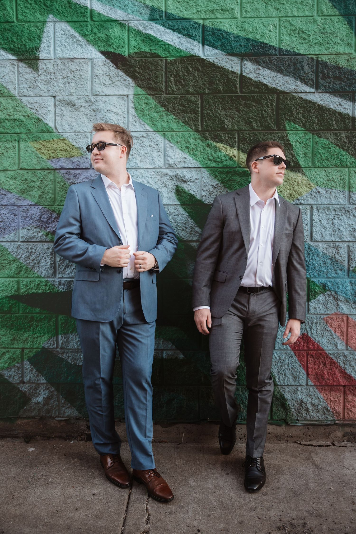Two men in suits wearing sunglasses posing against colorful graffiti brick wall.